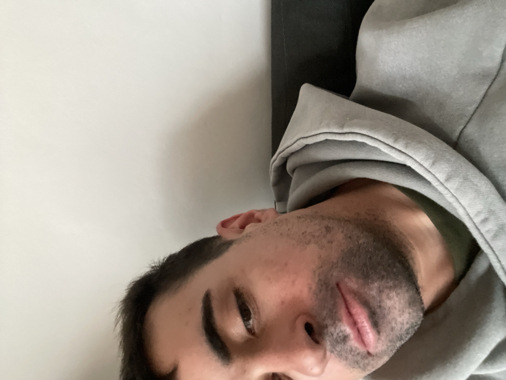 The image presents a close-up, angled shot of a man in a relaxed, possibly reclining, position indoors. His head is tilted to the right, occupying the lower right portion of the frame. He has dark hair, prominent dark eyebrows, and a short beard or heavy stubble covering his jawline and upper lip. His lips are slightly parted, and his dark eyes are partially open, looking towards the viewer with a neutral or calm expression.
The man is wearing a grey hooded sweatshirt, with the hood slightly pulled up, framing his face. A glimpse of a dark green shirt or garment is visible under the hoodie around his neck. The background consists of a plain white surface on the left side of the image, likely a wall. To the right, behind his shoulder and the grey hoodie, is a darker, indistinct surface, possibly a piece of furniture like a couch or bed. The lighting is soft, suggesting an indoor setting.
The scene conveys a casual and quiet moment of rest or relaxation. No specific activities are taking place beyond this posture. There are no visible location cues within the image to suggest Barcelona, Spain, and no text is discernible. The time of day and weather conditions cannot be determined from the photograph.