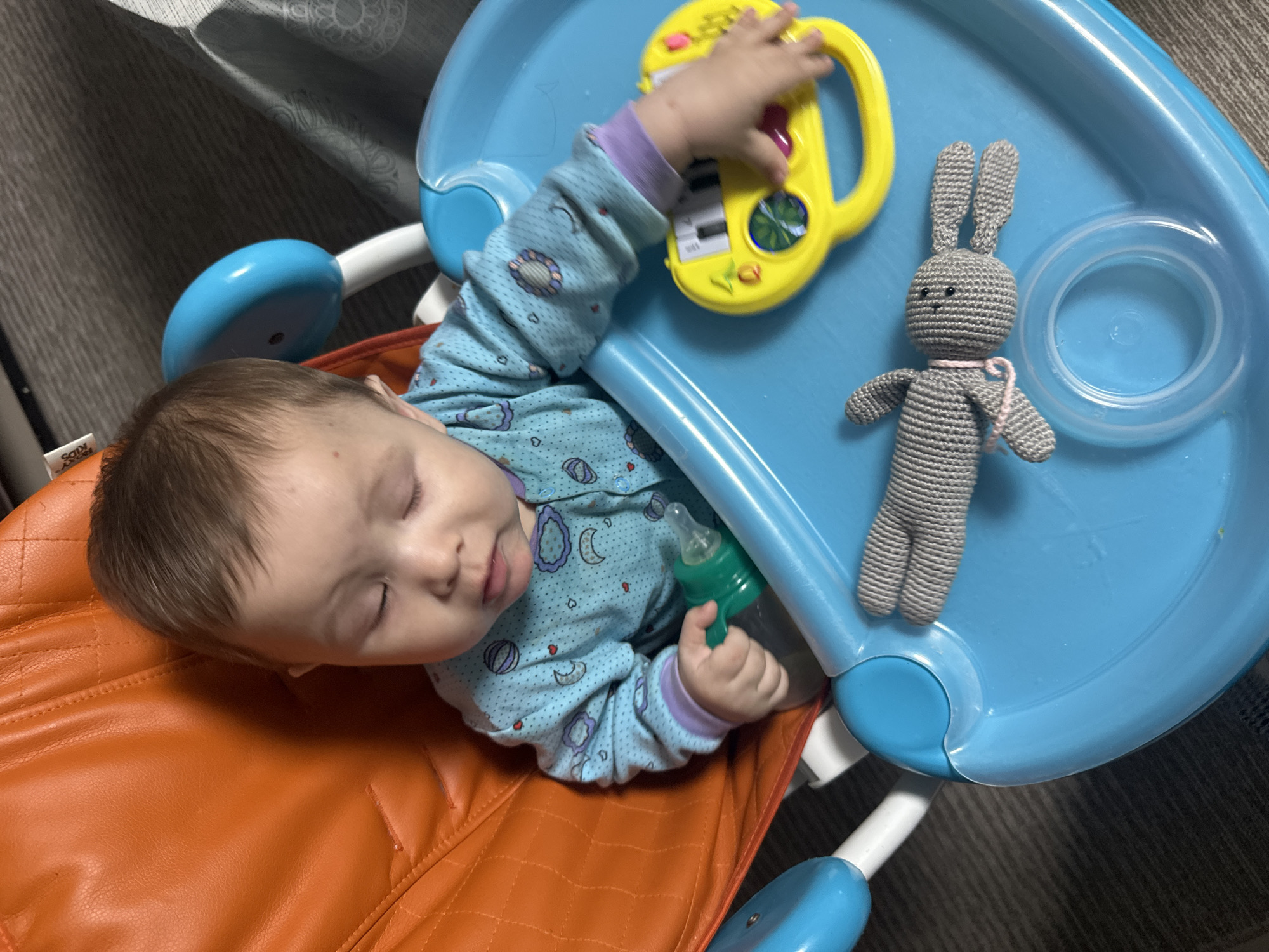 This image captures a baby sleeping peacefully in what appears to be a high chair or bouncer. The baby, lying on their left side, has light brown hair and closed eyes, suggesting deep slumber. They are dressed in a light blue long-sleeved onesie patterned with moons and various shapes.
The baby is positioned on an orange, quilted seat, where a white label bearing the word "KIDS" is partially visible on the left side. To the right of the baby is a blue plastic tray. On this tray, a yellow toy, resembling a miniature piano or musical activity board with colorful buttons, is placed, and the baby's left hand rests lightly upon it. Beside the toy, a gray crocheted bunny with a pink ribbon around its neck is also on the tray. The baby's right hand is loosely holding a green baby bottle or sippy cup.
The background shows an indoor setting, with a dark patterned curtain visible on the left side, and a dark carpet or rug beneath the high chair. The overall scene conveys a calm and quiet moment, likely during the daytime, with the baby resting amongst their playthings.