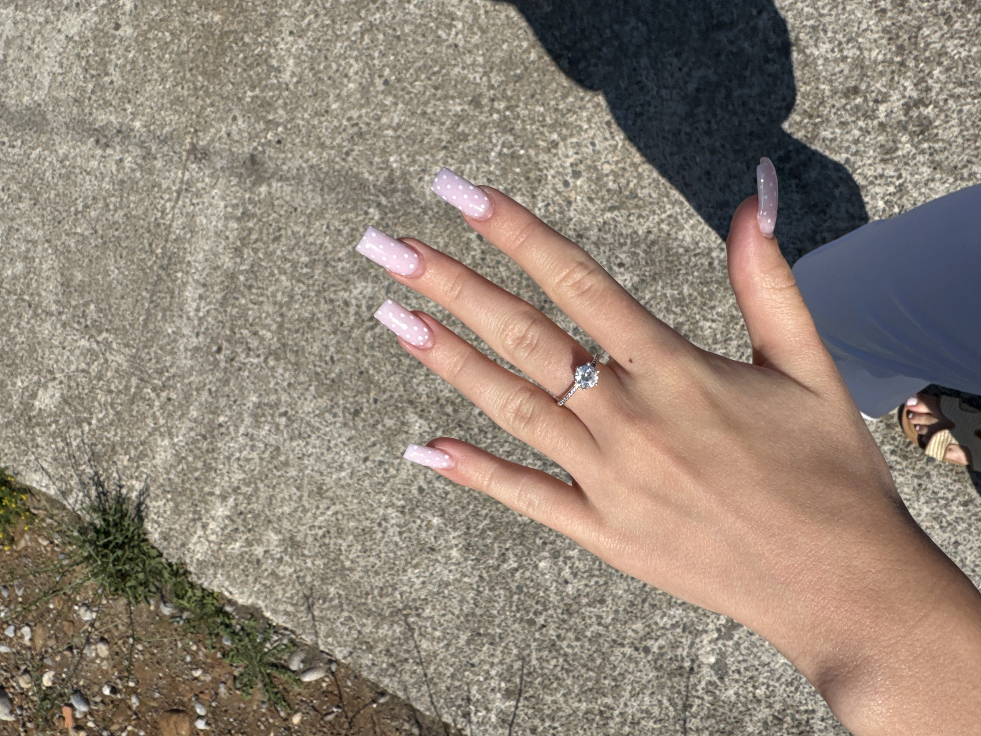 A person's left hand, adorned with light pink square-shaped acrylic nails featuring a white polka dot pattern, is prominently displayed against a textured concrete and gravel ground. A sparkling diamond ring is worn on the ring finger. The background shows a sliver of green vegetation and the shadow of the person creating a contrast on the concrete. The feet, wearing open-toed sandals, are visible at the bottom of the frame. The image is taken outdoors, under bright sunlight, suggesting a daytime scene. The location is indeterminate, as there are no discernible city landmarks or context.