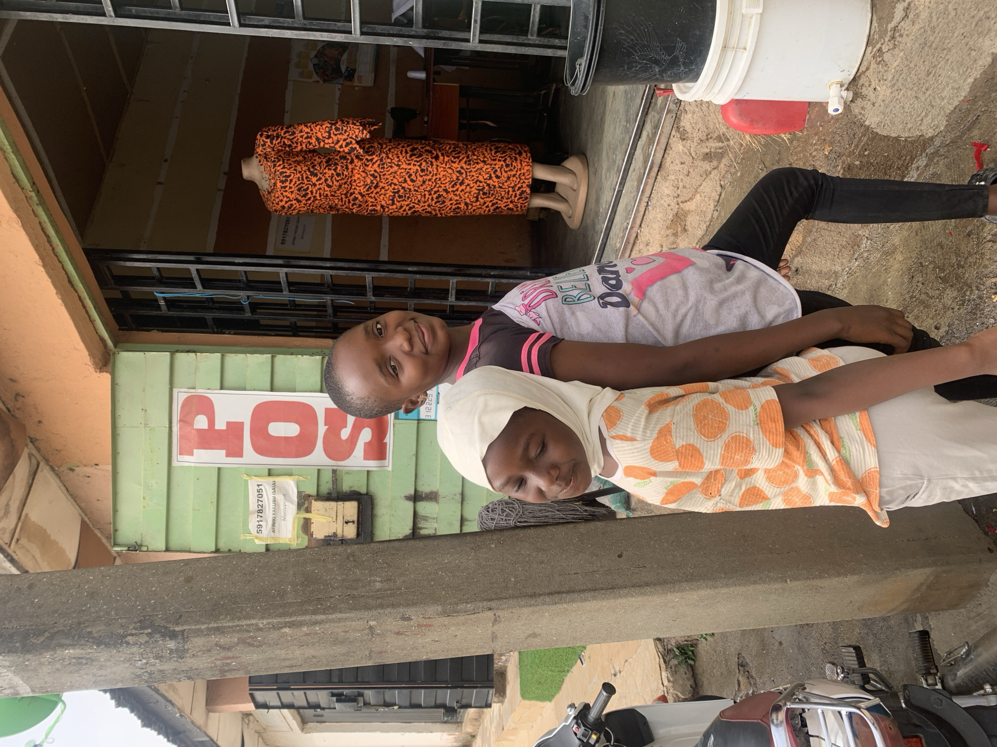 The image, taken in Jos, Nigeria, captures two young girls standing outdoors in front of a shop during daytime. In the foreground, a girl wearing a light-colored headscarf and an orange-and-white patterned top is positioned with her eyes closed or looking downwards. Beside her, another girl smiles towards the camera, wearing a grey t-shirt with pink and black accents and black trousers. Her arm is gently resting on the first girl's back, suggesting a close relationship.
They are situated behind a prominent concrete barrier or ledge that traverses the mid-ground. Behind them, the entrance to a shop is visible, secured by metal bars. Inside the shop, a mannequin displays an orange and black patterned dress. To the left of the shop entrance, a red sign, partially obscured, shows the letters "POS," likely indicating a Point of Sale service. Below this, a white paper taped to the wall displays text including "5917827051," "ARMIKI YAKUBU ISAIYAH," and "Moneypoint," appearing to be contact and business information.
The ground is concrete, with some areas appearing damp near the shop entrance, where a black bucket, a white bucket with a tap, and a red container are visible. A motorcycle or scooter is partially seen in the lower right corner. The natural light indicates it is daytime, and the overall scene depicts a casual street or commercial setting.