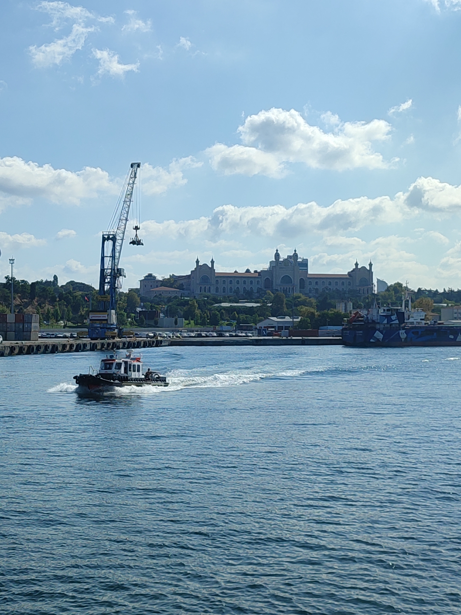 A wide shot shows a body of water in Istanbul, Turkey, on a clear, partly cloudy day. In the foreground, a small white and red boat speeds across the water, leaving a white wake. In the middle ground, a large blue crane is positioned over a dock where shipping containers are stacked. Behind the crane, a large, ornate building with domes and multiple levels sits on a hill. To the right of the building, a large blue cargo ship with a camouflage-like pattern is docked. The water is a deep blue, and the sky is a bright blue with scattered white clouds. The overall scene depicts a busy port or industrial area in Istanbul.