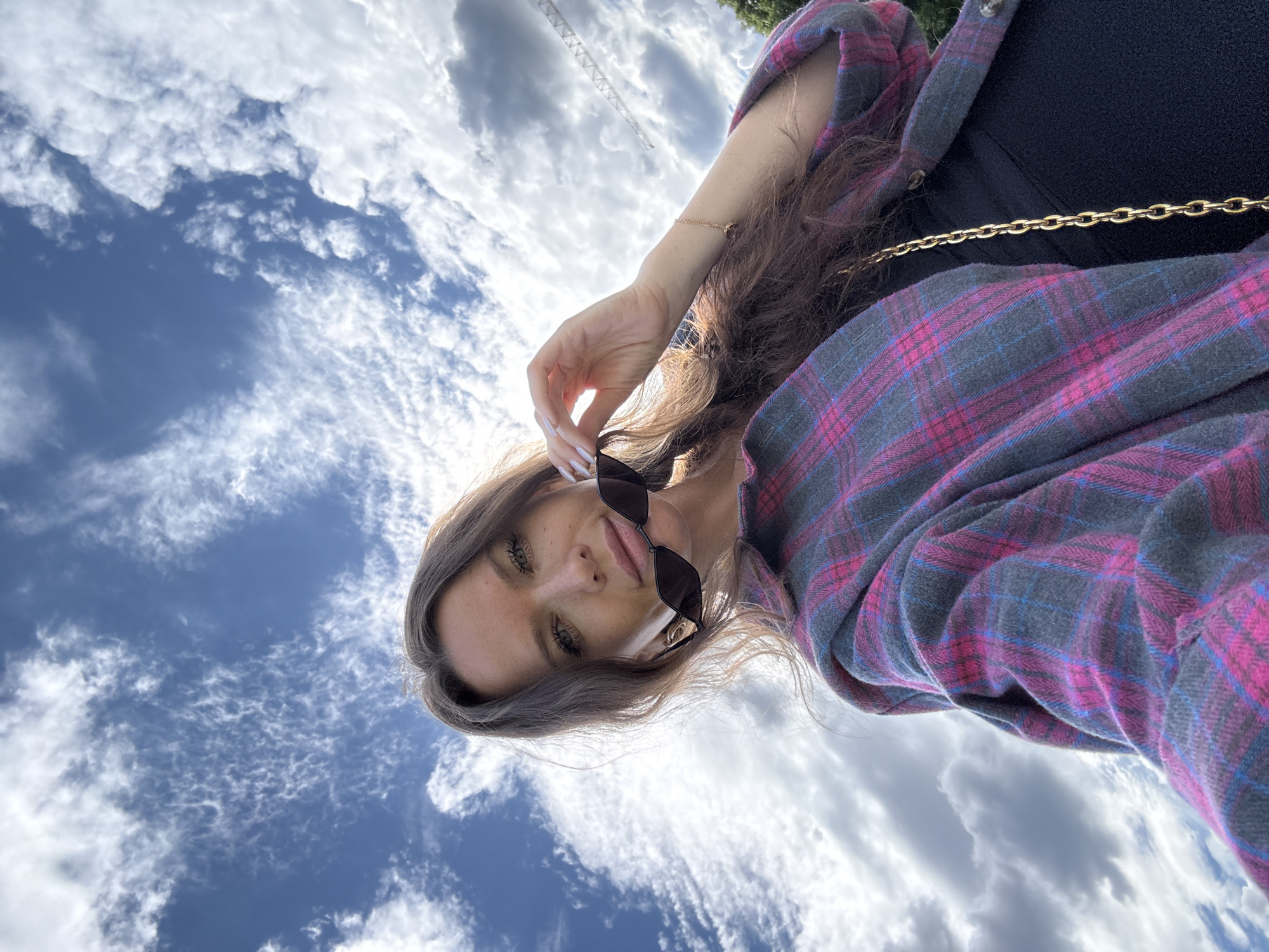 A woman with long brown hair and sunglasses is taking a selfie outdoors. She is wearing a pink and gray plaid shirt over a black top, and a gold chain purse strap is visible. The sky behind her is blue with white clouds. A construction crane is visible in the distance. The image is shot from a low angle, looking up at the woman.