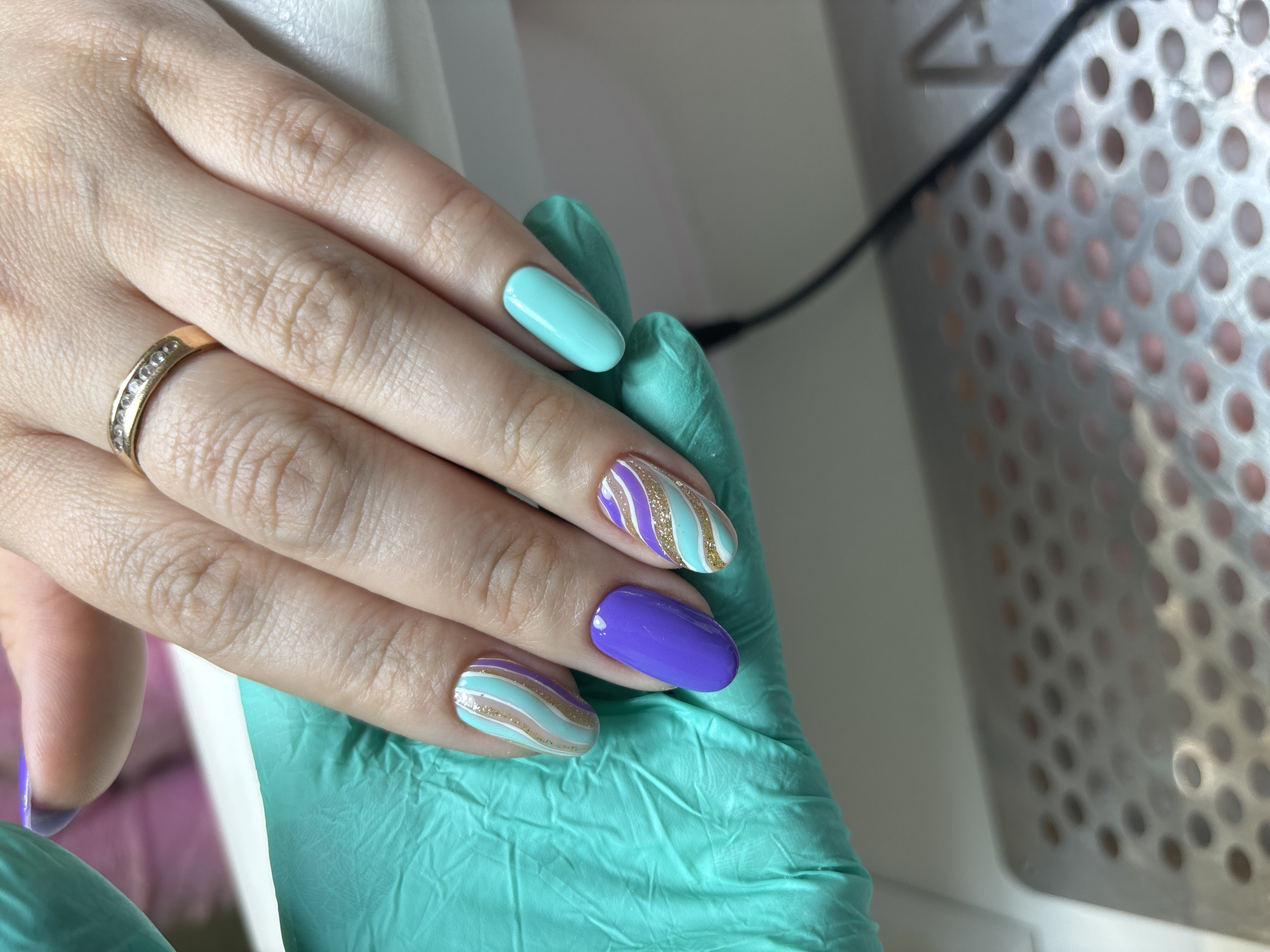 This close-up image features a person's left hand, adorned with a vibrant manicure, resting partially on a light blue-green nitrile glove. The back of the hand and fingers are visible, while the palm is covered by the glove. A gold-toned ring, set with a line of small, clear stones, is worn on the ring finger.
The fingernails display a creative and colorful design. The nail on the index finger is a solid light blue-green, closely matching the color of the glove. The ring finger nail is painted a solid, bright purple. The middle and pinky finger nails feature a striped pattern, alternating between light blue-green, purple, and shimmering gold glitter lines, creating a playful yet elegant look. The thumb nail, partially visible on the left, also appears to be a solid purple. All nails have a smooth, glossy finish and a well-maintained oval shape.
The background shows a light-colored surface, possibly a workbench or table. To the right, a metallic surface with numerous small perforations, characteristic of a nail dust collector or similar salon equipment, is visible, with a black cable traversing it. The lighting is bright and evenly distributed, highlighting the details of the manicure.
The scene suggests an indoor setting, likely a nail salon or a dedicated beauty space, showcasing the freshly applied nail art. No text is visible in the media file.
