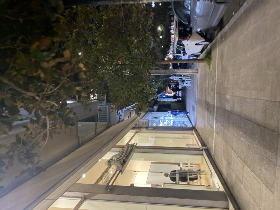 This media file captures a night-time street scene in Egaleo, Greece. The perspective is looking down a sidewalk lined with shops, trees, and parked cars, suggesting an urban or commercial area.
On the left side of the image, a modern building with large glass windows houses a brightly lit shop. Inside, a person wearing dark clothing is seated at a table or desk. Attached to the building's exterior, above the lower windows, is a metallic, ladder-like structure, possibly part of a window cleaning system or an awning mechanism. An extended awning provides shelter over the sidewalk immediately in front of this shop. Further down the street, another shop is visible with a strong blue light emanating from its interior, and a person in a blue shirt is standing outside it.
The right side of the image features a wide sidewalk paved with light grey and darker grey square tiles. Parallel to the sidewalk, several cars are parked along the curb, including a silver sedan and a white hatchback. Trees with dark green foliage line the street between the sidewalk and the parked vehicles, their leaves partially obscuring the background. Streetlights are visible further down, illuminating the scene. In the distance, more buildings and what appears to be an outdoor seating area for a cafe or restaurant can be discerned.
The time of day is clearly night or late evening, as indicated by the dark sky and the reliance on artificial lighting from the shops and streetlights. No specific activity beyond people standing and walking, and one person inside a shop, is clearly discernible. A small white letter 'D' on a red background is visible on a sign to the left of the initial shop's entrance.