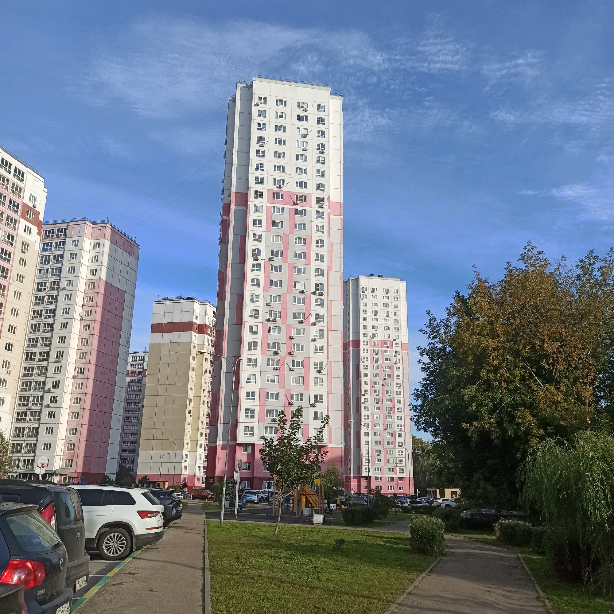 This is a ground-level, eye-level, wide shot photograph taken on a sunny day with a clear blue sky dotted with wispy white clouds. The scene depicts a residential area in the city of Nizhny Novgorod, Russia, characterized by tall, multi-story apartment buildings.
The foreground shows a paved walkway bordering a patch of green grass. Several cars are parked along the edge of the walkway. The cars are of various models and colors, including a dark hatchback in the immediate left foreground, a white SUV, and other dark-colored vehicles further back.
In the middle ground, a children's playground with a yellow and red slide structure is visible, surrounded by grass and some bushes. A young tree stands in front of the playground.
The background is dominated by several large apartment buildings. The buildings are tall and rectangular, with numerous windows. They feature a color scheme of white, light gray, and pink panels. Some buildings have balconies or air conditioning units visible on their facades. In the distance, a denser cluster of similar buildings can be seen.
To the right of the frame, a large tree with green and brown foliage stands prominently, its branches extending towards the buildings and the sky. A weeping willow-like tree with long, drooping branches is also visible further to the right. The overall impression is of a modern, urban residential complex under a bright, cloud-streaked sky.
