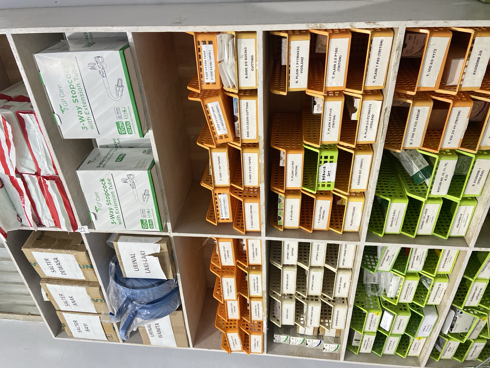 The image shows a storage shelf filled with medical supplies. The shelf is divided into multiple compartments, each holding boxes or containers of various medical items. The items appear to be organized by type and possibly by brand. Many of the containers have labels with text in English, some of which are partially visible. Examples of visible text include "GP Care," "3-Way Stopcock with Extension Tube," "CUTTING," "VIGILENZ," "TRITON," "JARUM," and product codes like "B.SIDE 3-0 5266." There are also labels in Indonesian, such as "URINAL LAKI-LAKI," "URINAL WANITA," "SALTER DEWASA," "SALTER ANAK," and "SALTER BAYI." The setting appears to be a medical storage area or pharmacy, likely in Indonesia given the Indonesian text. The lighting suggests an indoor environment. There are no people visible in the image, and the focus is solely on the organized medical supplies.
