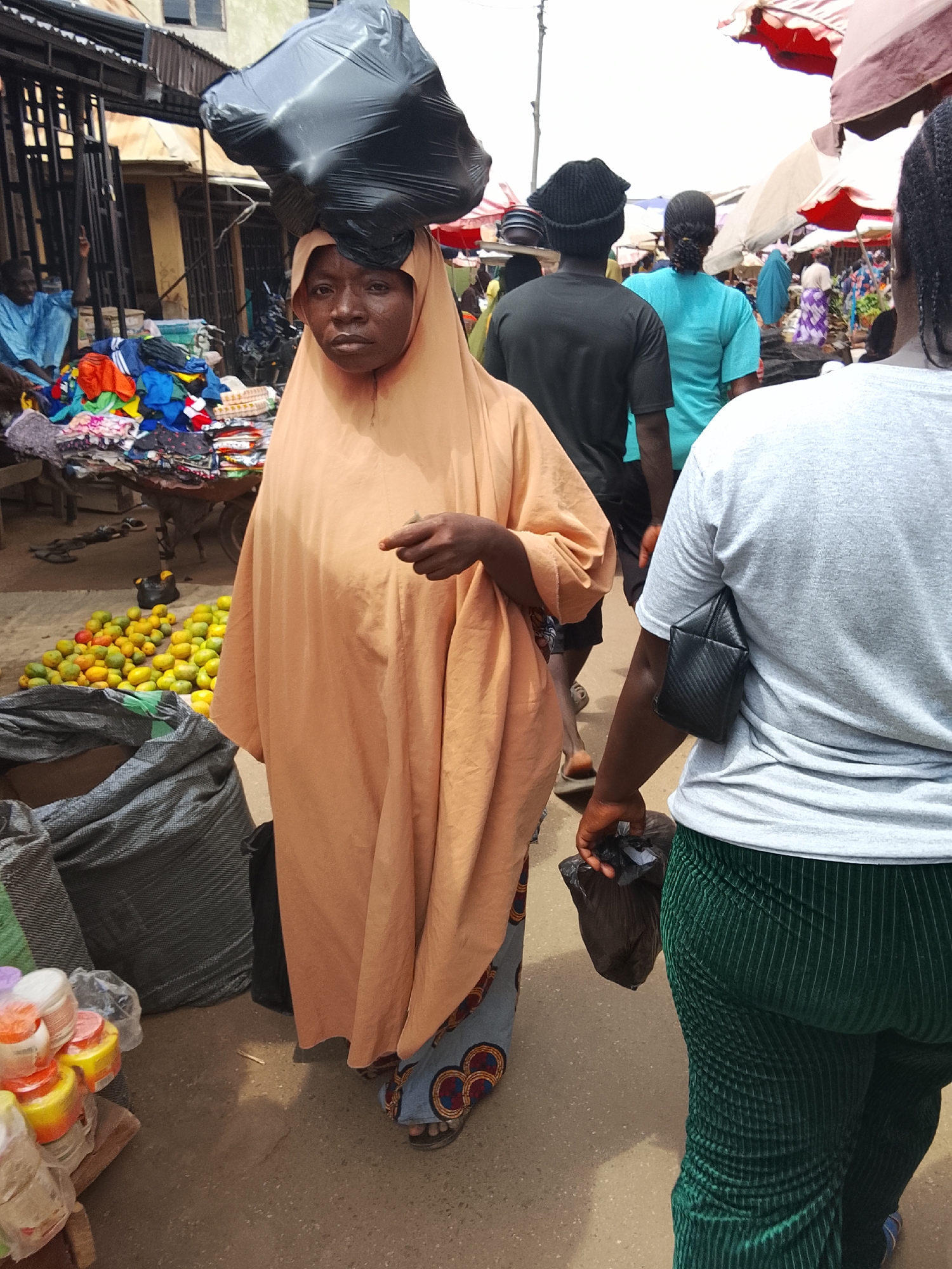 In a bustling outdoor market in an unidentified city, an adult woman with dark skin carries a large black bag on her head. She wears a light-orange headscarf and long robe over a patterned fabric skirt. She holds her left hand out slightly. The market surrounding her includes tables of colorful clothing and a pile of yellow and green fruit. Another woman in a gray shirt and green pants walks beside her, carrying a black plastic bag. Other people are seen in the background, along with various market stalls under colorful parasols.