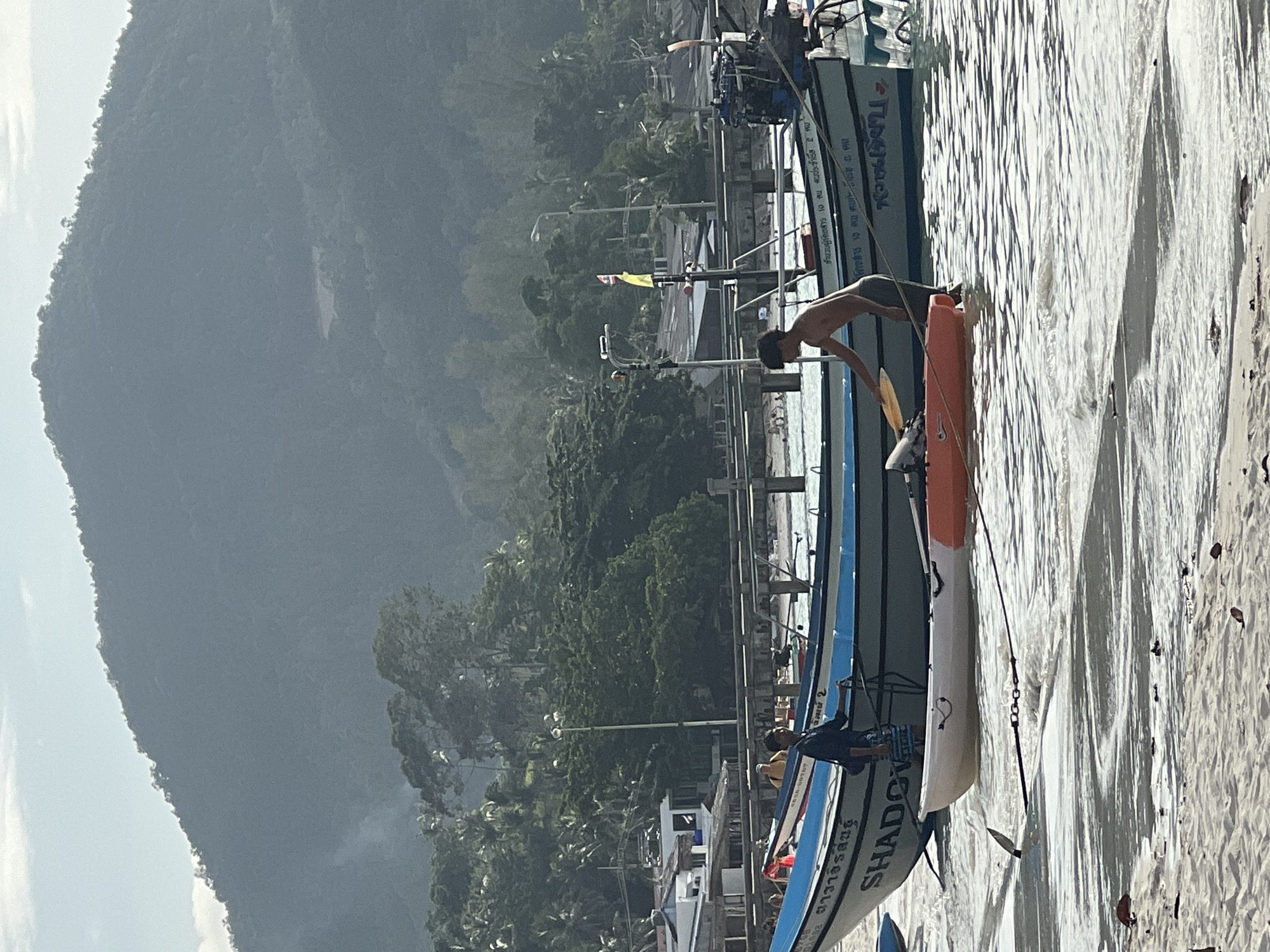 The image depicts a beach scene with several boats and two people. In the foreground, a sandy beach is partially submerged by shallow, rippling water. An orange and white kayak is floating near the shore, tethered by a rope to an anchor. To the left of the kayak, a dark object, possibly a piece of debris, is visible in the water.
Further out, a larger blue and white boat is anchored. A young man, shirtless and wearing dark shorts, is standing in the water next to this boat, holding a yellow paddle. He appears to be interacting with the kayak. Another person, a man in a yellow shirt, is standing on the deck of the blue and white boat.
Behind the boats, a wooden pier extends into the water, with several structures and a light pole visible on it. The background is dominated by a lush green, tree-covered mountainside shrouded in a light haze. The sky above is pale blue with some wispy clouds. The overall lighting suggests it is daytime, possibly late morning or early afternoon. The scene appears to be a coastal area, likely in a tropical or subtropical region, given the vegetation and boats.