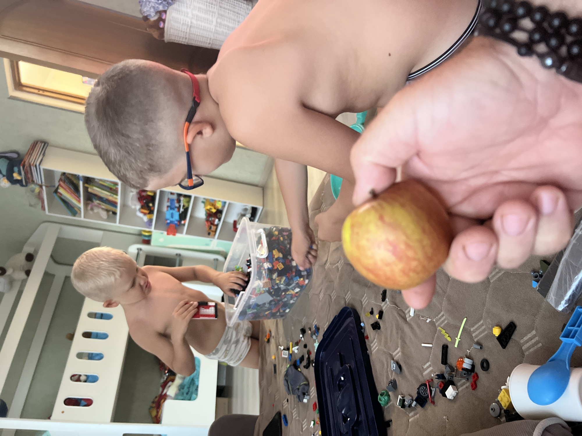 **Scene Description:** An indoor residential scene, likely a child's play area, is captured from a low, close perspective. The primary focus is on two young male subjects engaged in playing with plastic building blocks. A third, adult subject's hand is visible in the immediate foreground, holding a fruit. **People:** 1. **Subject 1 (Left):** A male child, estimated age 4-6 years, with light blonde, short-cut hair. He is shirtless and wears light-colored boxer briefs with a subtle, dark patterned print (e.g., elephants). He stands or kneels next to a clear plastic container of building blocks, looking down and reaching into it with his right hand. His left hand holds a small red rectangular object. 2. **Subject 2 (Right):** A male child, estimated age 6-9 years, with short, light brown hair styled in a buzz cut. He is shirtless and wears dark-colored underwear with a white stripe along the waistband. He wears blue and orange framed eyeglasses with a red strap securing them around his head. His back is partially turned to the camera as he leans forward, his right arm extended towards the building blocks on the table surface. 3. **Subject 3 (Foreground):** A human right hand is visible in the lower right corner, holding a small, round fruit with mottled reddish-yellow skin, resembling an apple or plum. A dark, beaded bracelet is present on the wrist. **Objects and Environment:** * **Playing Surface:** The children are interacting with numerous colorful plastic building blocks (resembling Lego) spread across a brown fabric surface, likely a tablecloth, exhibiting a repetitive, lighter brown rectangular or square pattern. A large, clear plastic storage container, approximately one-third full of these blocks, rests on this surface. * **Background Left:** A white wooden bunk bed is visible, with the lower bunk containing colorful textiles and bedding. The upper bunk's side rail features multiple vertical rectangular cutouts, each containing a blue insert. A white open shelving unit to the left of the bunk bed displays various books and toys. * **Background Right:** A second white open shelving unit displays books, small toy vehicles, and stuffed animals. Adjacent to this, a white woven basket filled with soft items is present. A light-colored doorway is visible in the upper right, with a warm, yellow light emanating from the space beyond it. * **Foreground and Mid-ground Objects:** A dark blue plastic tray or lid, a white plastic cup containing a blue plastic spoon,