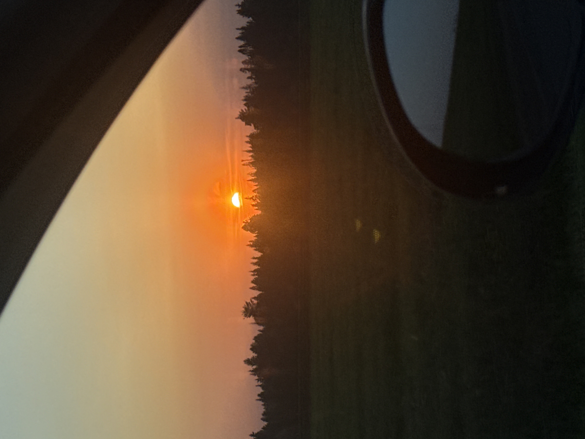 The image shows a sunset through a car window. The sun is positioned low on the horizon, obscured by a line of trees. The sky near the sun is a gradient of orange and yellow. A car's side mirror is visible in the upper right corner. The overall scene appears to be captured from inside a moving vehicle. There is no visible information related to the location within the city of Kamennoye, Russia.