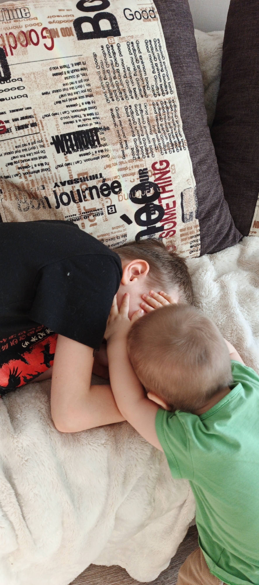 Two young children are lying on a beige plush blanket. One child, wearing a black t-shirt with a red and black design on the sleeve, is mostly obscured. The other child, wearing a green t-shirt, has his hands on the face of the first child. The setting is indoors. Behind the children are two pillows, one with a print featuring words like "Good" and "Goodbye" and another that is dark brown. The location is Chaikovsky, Russia.
