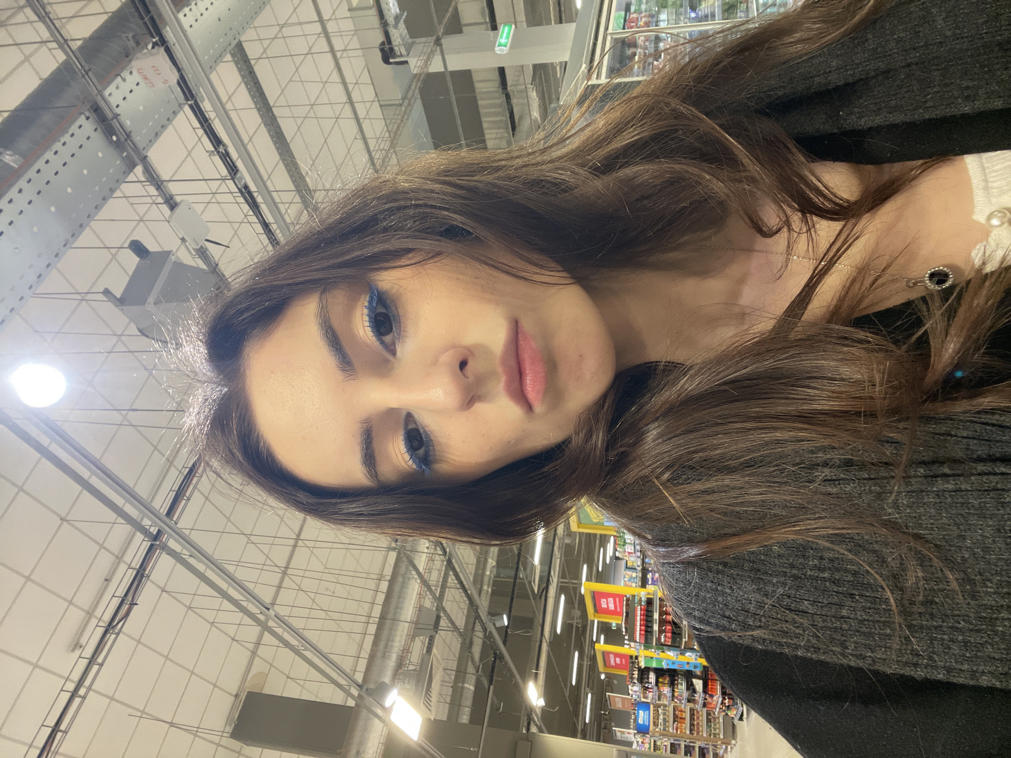A young woman is looking into the camera, presumably taking a selfie. She has long brown hair and is wearing blue eyeliner. She appears to be in a large retail store with shelves of products visible in the background. The location could be Moscow, Russia. The background includes ceiling structures and lighting typical of large commercial spaces.