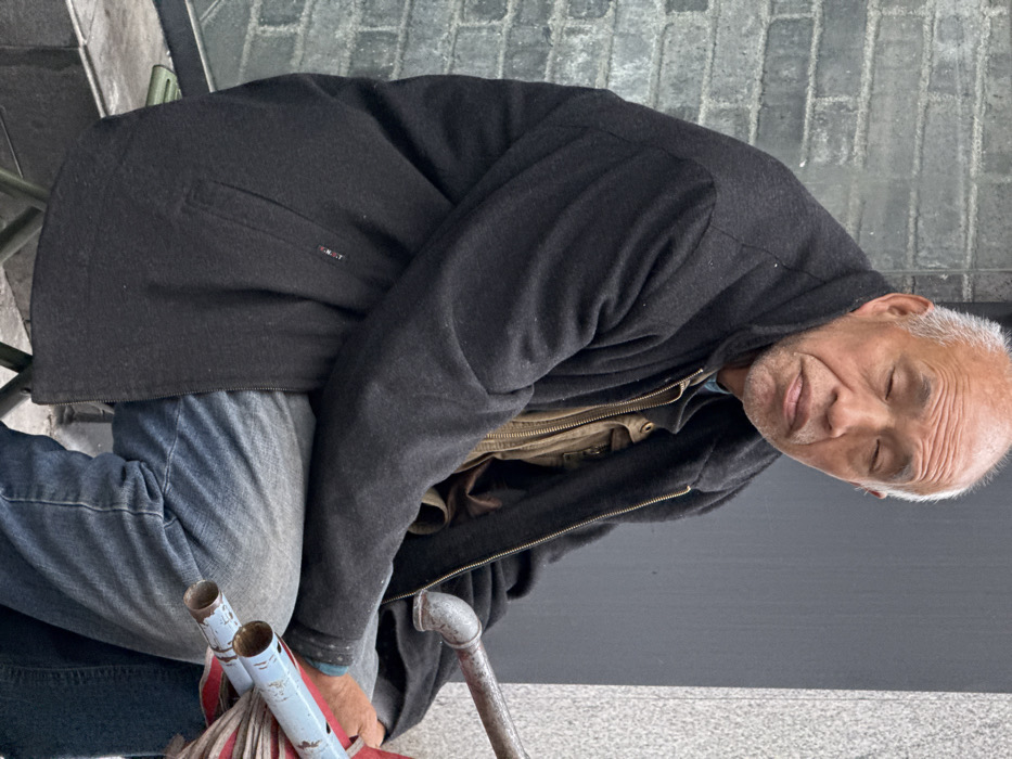 An East Asian male, appearing to be elderly, with a bald head and short white hair at the temples, is depicted seated outdoors in an urban environment. His eyes are closed, and his facial expression is neutral. He is wearing a dark charcoal-colored, long-sleeved jacket zipped over a lighter brown or tan garment, possibly a vest or jacket, with a blue collar visible underneath. He wears blue denim jeans.
He is seated on an unseen object, with parts of what appears to be a light green or grey metal frame visible beneath him to his lower left. To his right, partially visible, are several objects with metal piping. These items consist of a bundle of red material and at least two blue cylindrical metal pipes, suggesting a folded or disassembled apparatus, possibly a street vendor's stall component or a large umbrella.
The background features two distinct wall textures: a dark grey brick wall to the viewer's left and a smooth, light grey building facade to the viewer's right. The scene is consistent with a street or sidewalk setting within an urban area, such as Yuzhong District, China.