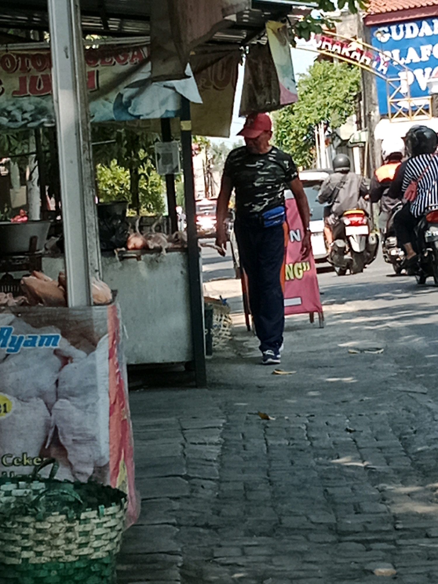 The image depicts a street scene in Banaran, Indonesia, characterized by a sidewalk made of grey bricks and an asphalt road. In the foreground, a person wearing a camouflage t-shirt, a red cap, and dark pants with a blue fanny pack is walking away from the camera. To the left, a market stall is visible, with various items including sacks of what appears to be chicken, a basket, and signs. One sign has text including "Ayam," which means chicken in Indonesian. In the background, several people are seen on motorcycles. One motorcycle is ridden by a person wearing a grey top and a helmet. Another motorcycle carries two people, one in an orange top and another in a black and white striped shirt and helmet. Behind the people on motorcycles, a blue building with white and red text, likely a shop or business, is visible. The environment suggests a busy marketplace area in a city. The lighting indicates it is daytime, with shadows cast on the ground.