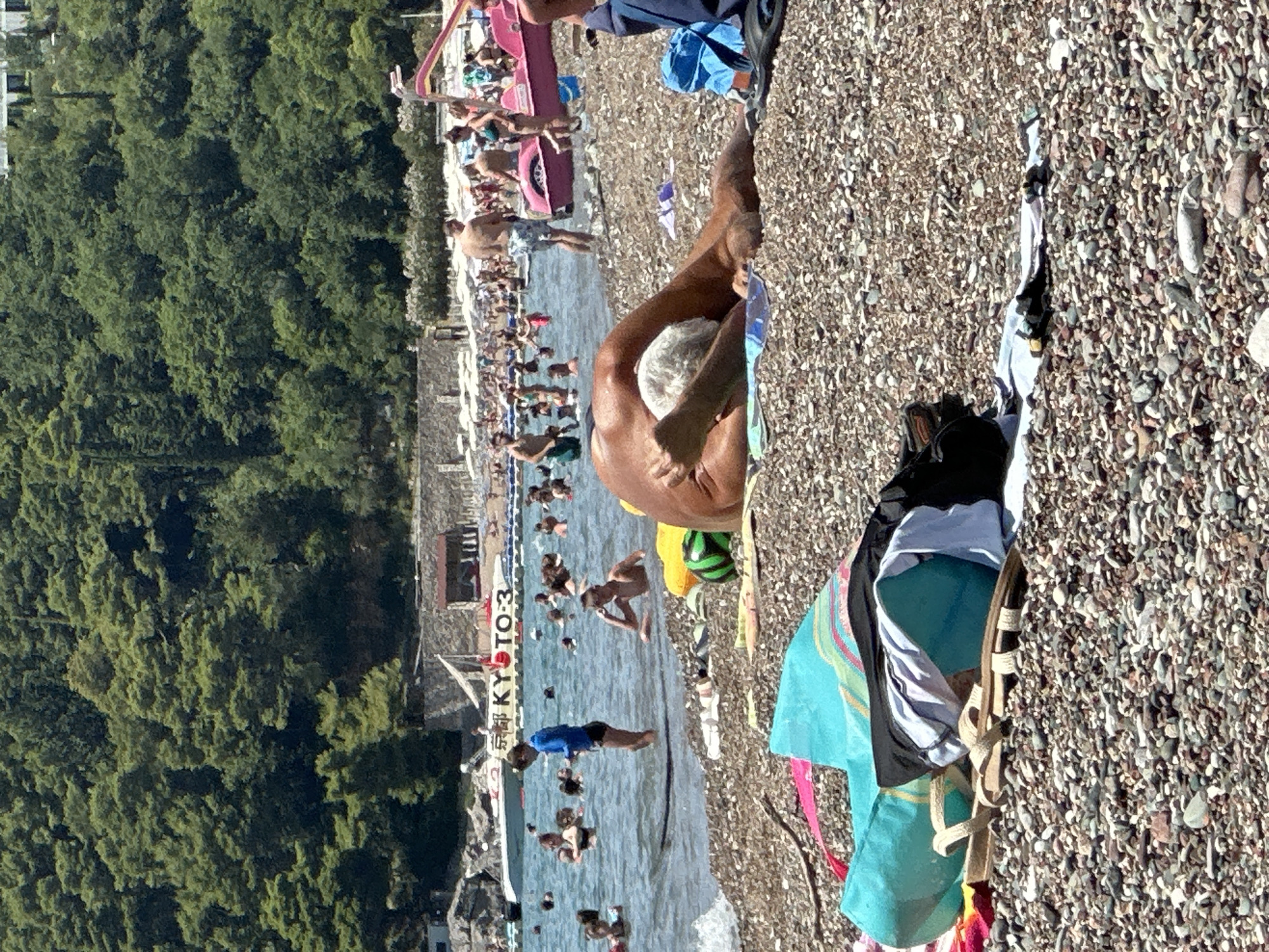 The image depicts a crowded beach scene in Boreti, Montenegro, on a sunny day. In the foreground, a person is lying on their stomach on the pebbled shore, with their back to the camera, sunbathing. Their face is obscured, but their white hair is visible. Beach towels and bags are scattered around the person.
In the mid-ground, the shallow, clear water is filled with people swimming and wading. A small boat with the text "KYOTO-3" is visible on the water. Further back, a stone wall and a line of beach umbrellas indicate the edge of the beach.
The background is dominated by a dense, green forest covering the hillside, typical of the Montenegrin coast. The overall atmosphere is one of a bustling summer day at the beach, with numerous individuals enjoying the water and sun.