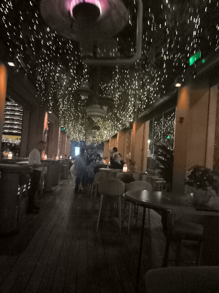 This low-light photograph captures the interior of a modern restaurant or bar during the evening. The most striking feature is the ceiling, densely covered with numerous small, bright string lights that create a sparkling, "starry night" ambiance. Several larger, dark, bowl-shaped pendant lights also hang from above. In the foreground, a prominent dark fixture, possibly an industrial-style heater or lamp, emits a distinct purple-pink glow from its core.
The setting includes a dark wooden floor and various seating arrangements. On the left, a bar area is visible with shelves displaying bottles, and a man in a light-colored shirt stands near the counter. Numerous patrons are scattered throughout the mid-ground, seated or standing around round tables with upholstered chairs and stools, engaged in socializing. Small, warm-toned lamps or candles provide accent lighting on the tables.
The right side of the space features large glass panels or windows, through which faint outdoor lights and some plants are discernible, suggesting a view to an exterior area or a covered patio. Warm, orange-toned architectural pillars or wall sections line this side. Small green rectangular lights are mounted on the upper wall or ceiling, possibly serving as indicators or exit signs. The overall atmosphere is relaxed and social, characteristic of an evening gathering in a contemporary establishment. No discernible text is visible in the image.