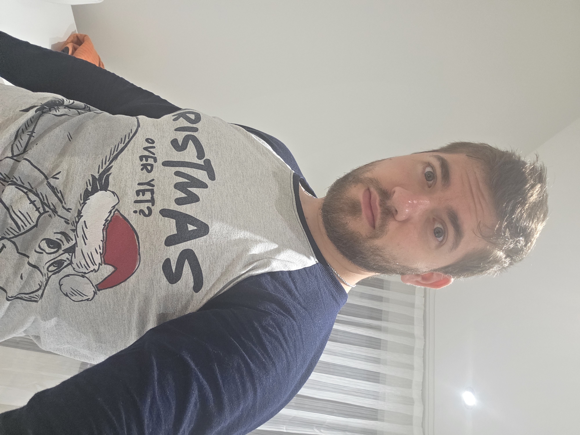 A man is wearing a grey, long-sleeved shirt with blue sleeves. The shirt has a cartoon design of a character wearing a Santa hat, along with the text "RISTMAS OVER YET?". The man has short brown hair and a beard. He is standing in front of blinds. The location is unspecified, and there is no information to indicate the location is Yagodovo, Bulgaria.