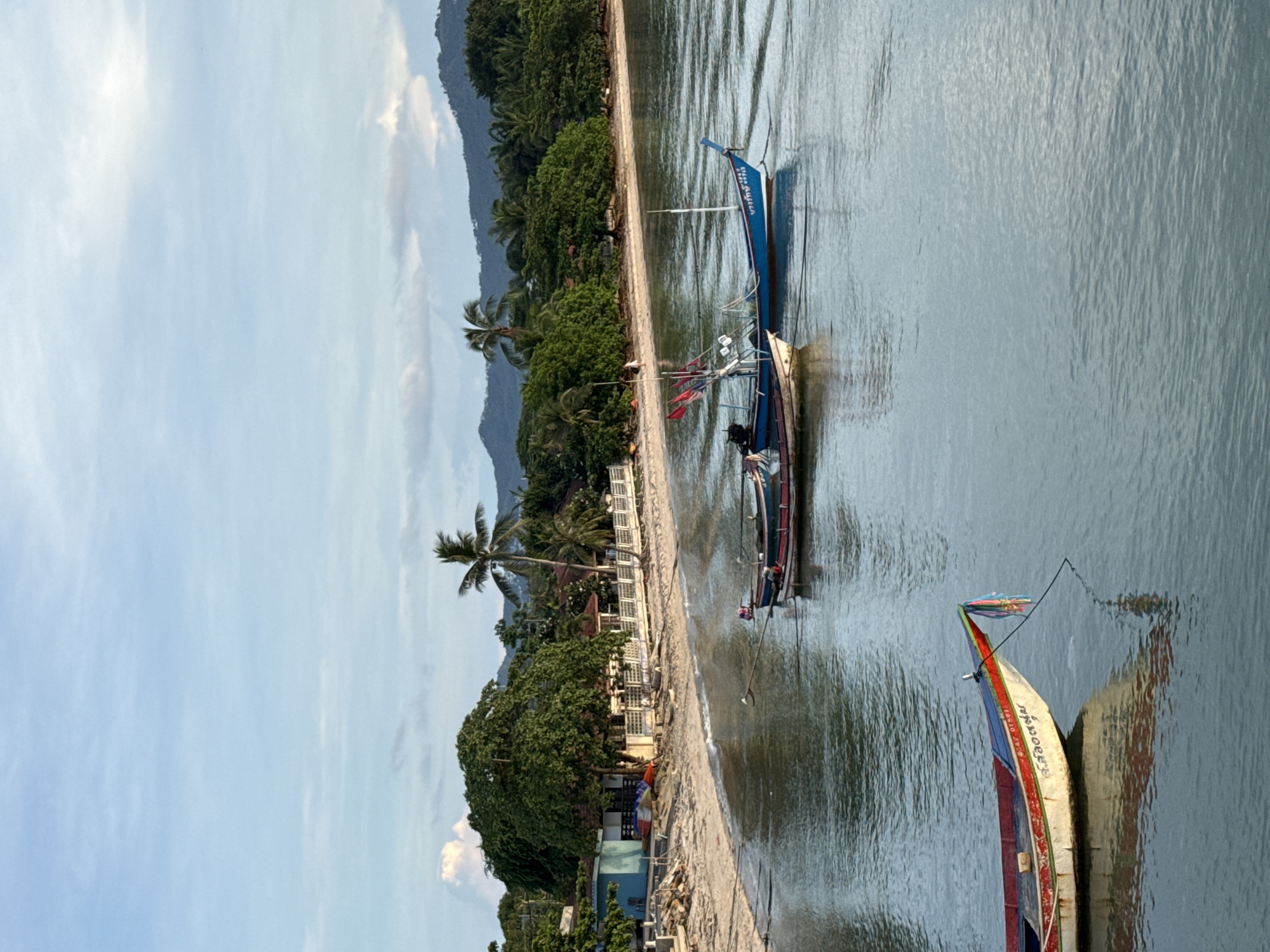 The image depicts a serene coastal scene at Baan Plai Laem, Thailand, during what appears to be early morning or late afternoon, indicated by the soft lighting and diffused sky. In the foreground, a colorful fishing boat, predominantly white with red and green accents, rests on the calm, clear water. Its reflection is visible below. In the middle ground, two more fishing boats are anchored closer to the shore. One is blue and white with Thai writing on its side, and the other is smaller and darker, adorned with red flags.
The shoreline features a sandy beach leading up to lush green trees and vegetation. Several buildings, likely residences or small businesses, are nestled amongst the trees, characterized by their tiled roofs and simple architecture, with one distinctly blue structure on the left. A white fence runs along the edge of the property facing the beach. In the background, a range of verdant, rolling hills stretches across the horizon, under a sky painted with pale blue and scattered, wispy clouds. A single palm tree stands prominently on the beach, adding to the tropical ambiance. The overall impression is one of peaceful coexistence between human activity and the natural environment.