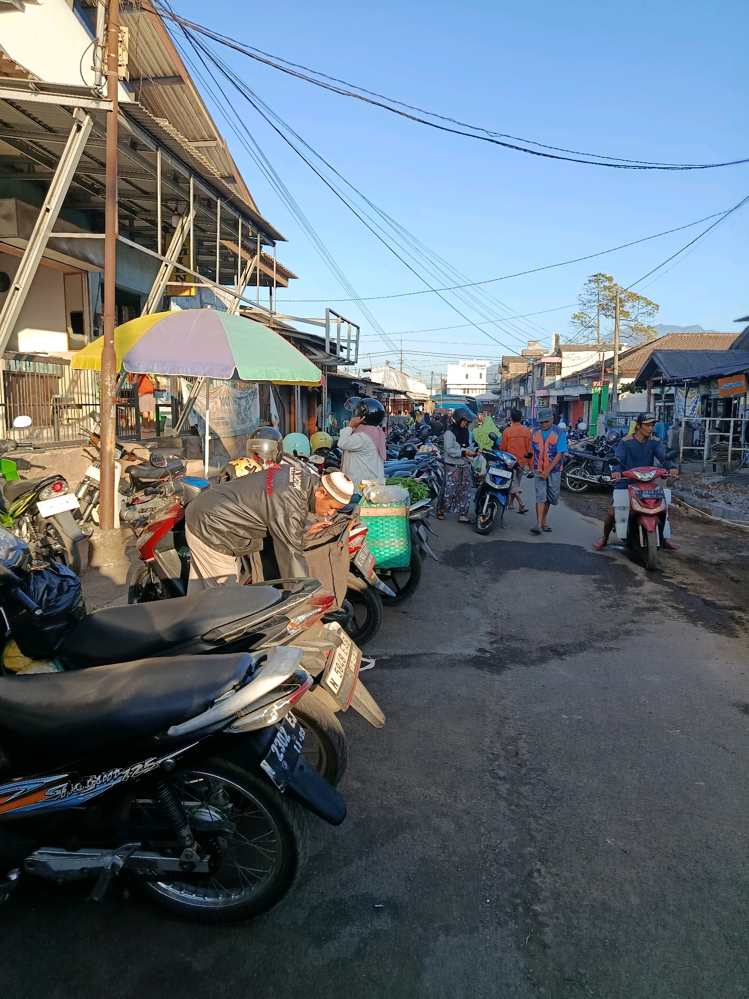 The image depicts a bustling street scene in Bululawang, Indonesia, likely in the morning given the clear blue sky and soft lighting. The street is lined with various shops and stalls, many with corrugated metal roofs. A prominent element is the large number of parked motorcycles, suggesting a busy market or transportation hub.
In the foreground, several motorcycles are parked on the asphalt road. One motorcycle in the lower left has a license plate that appears to read "N 2302 EL". To the right of this, a man wearing a black jacket and a white skullcap is bent over a motorcycle, appearing to be engaged in some activity. He is smoking a cigarette. Behind him, a colorful umbrella shades a small area.
Further down the street, more motorcycles are parked, and people are moving about. A man in an orange vest and blue shirt is walking, holding a bag. Another man is riding a red motorcycle, wearing a blue shirt and shorts. Several other individuals are visible, some walking, some standing, and one woman is wearing a headscarf and a pink top. A green plastic basket filled with produce sits on a motorcycle.
The environment is a typical street in a town or city, with buildings on either side, overhead power lines, and a slightly uneven road surface. The presence of numerous motorcycles indicates a common mode of transportation and possibly a local market day. The overall impression is one of everyday activity and commerce.