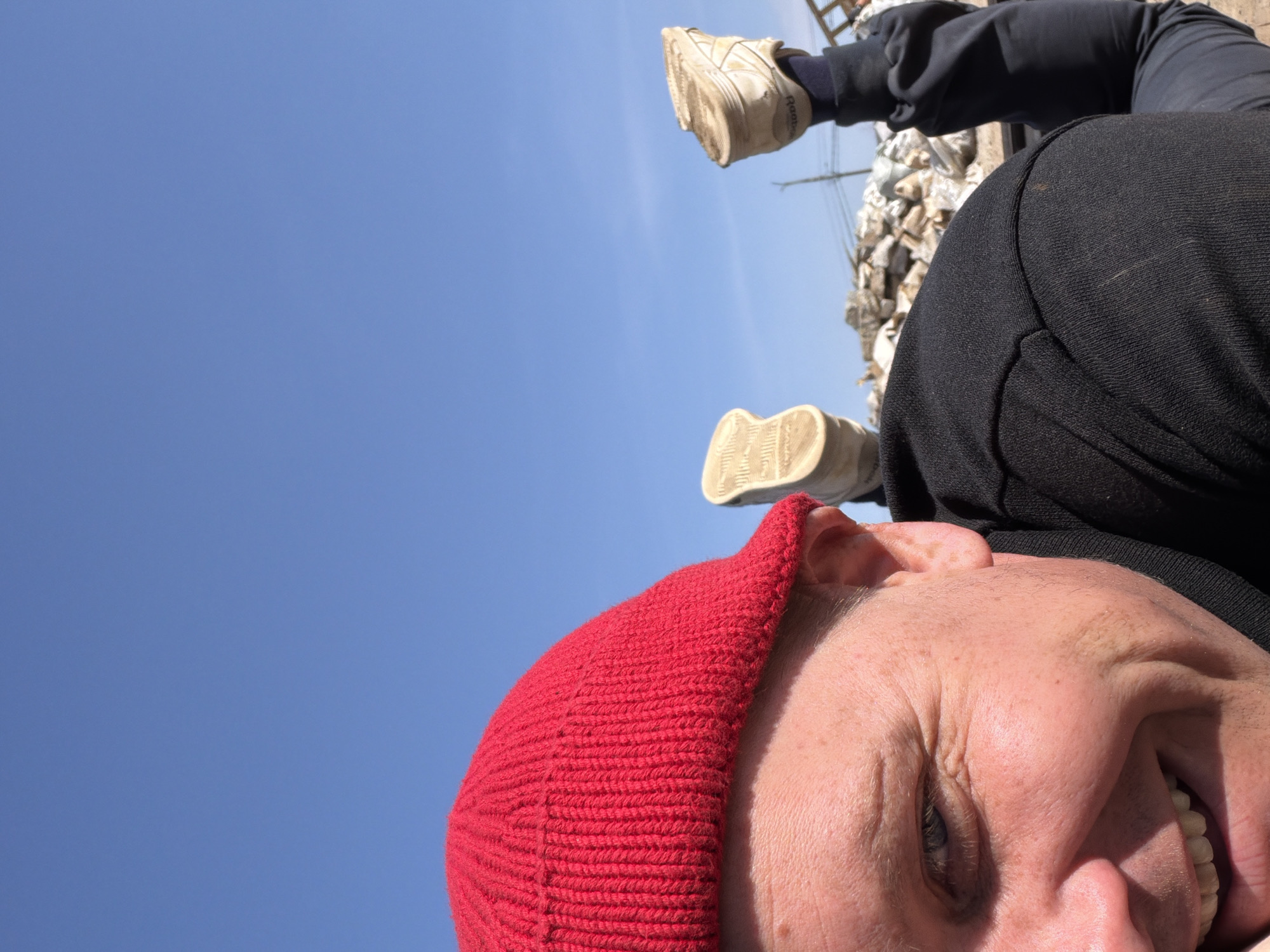 This image, taken in Chaikovsky, Russia, features a person captured from a low, rotated angle, suggesting they are lying down or in a highly reclined position looking upwards. The subject's face, partially visible in the foreground, wears a vibrant red knit beanie and has a joyful expression, with their mouth open in a wide smile or laugh, revealing teeth. Freckles are visible on their sunlit skin.
Above and behind the person's head, their legs are raised, with two feet clad in light-colored athletic shoes visible against a clear blue sky. The brand "Reebok" is discernible on the side of one shoe. The person is also wearing a black long-sleeved top and dark trousers.
In the background, beyond the person's legs, there's a pile of what appears to be rubble, bricks, or debris, along with a wooden or metal structure resembling a ladder. Faint utility lines are also visible against the bright sky. The scene is illuminated by clear, direct sunlight, indicating a daytime setting with good weather. The overall impression is one of happiness and playfulness in an outdoor environment.