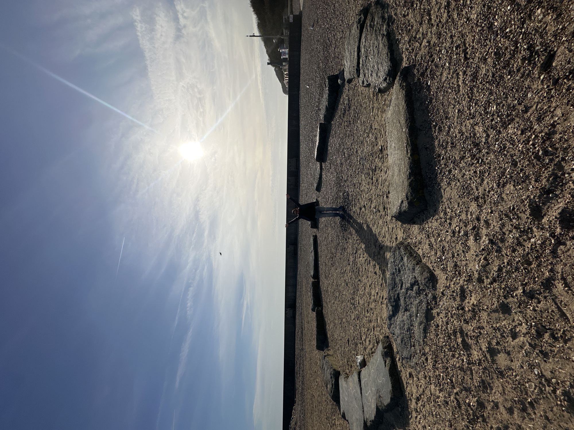 A rotated, wide outdoor shot shows a sunny day at the coast of Folkestone and Hythe District, United Kingdom. A person stands with their arms outstretched, casting a shadow onto the sandy ground. The ground is lined with dark, irregularly shaped rocks. A low concrete wall separates the sandy area from a road, beyond which a cluster of buildings can be seen in the distance. The sky is partly cloudy, with the sun shining brightly. A faint contrail from an aircraft is visible in the sky. A small bird is flying near the person.
