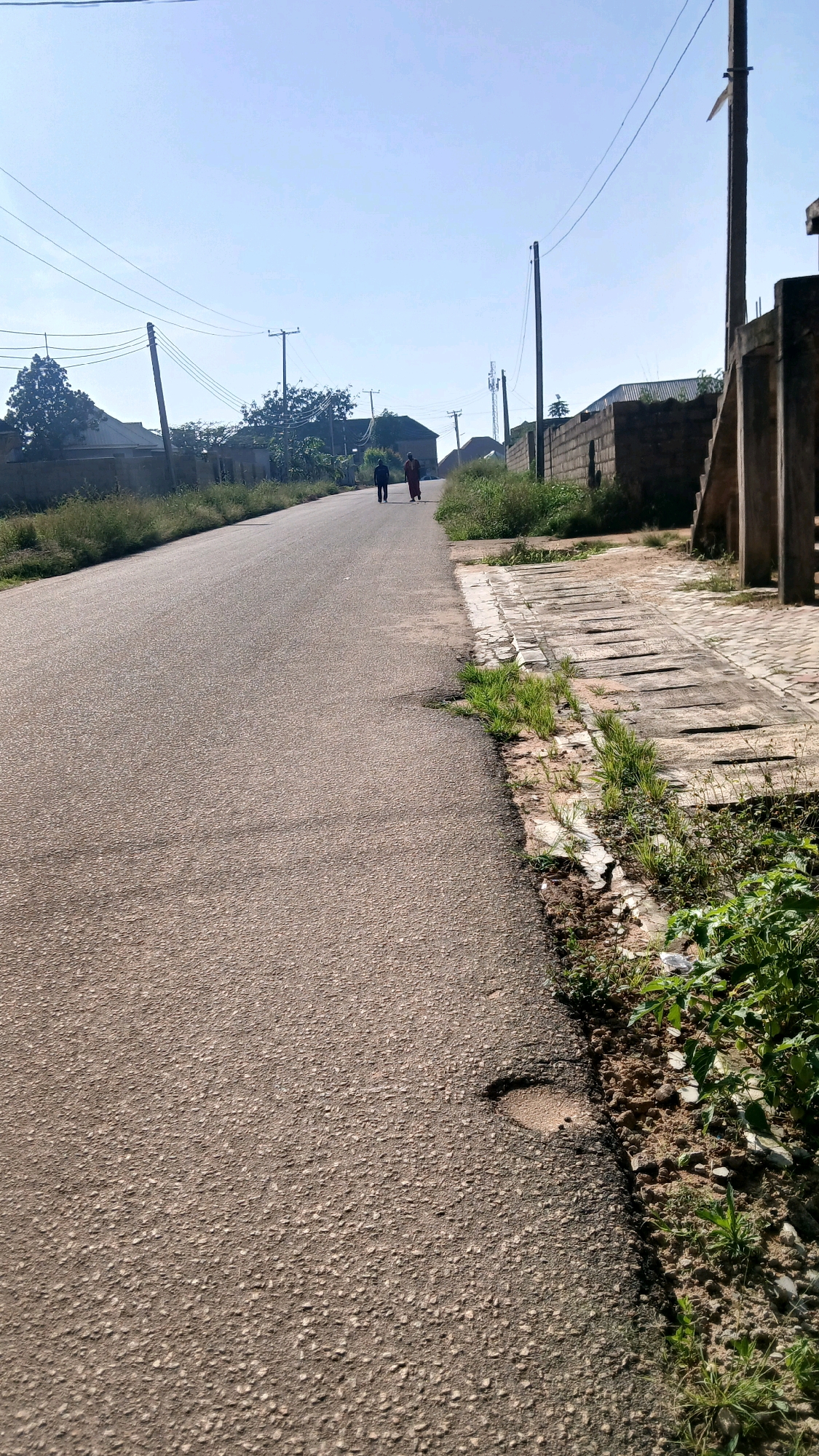 A long shot taken on a sunny day depicts a paved road in Gura Top, Nigeria, with two figures walking away from the camera. The road is asphalt with some cracks and a pothole visible on the right side. To the right of the road is a concrete drainage channel with overgrown grass along its edges. A stone wall and a concrete structure with stairs are also visible on the right. On the left side of the road, there are houses, trees, and utility poles with overhead wires. The sky is clear and blue.