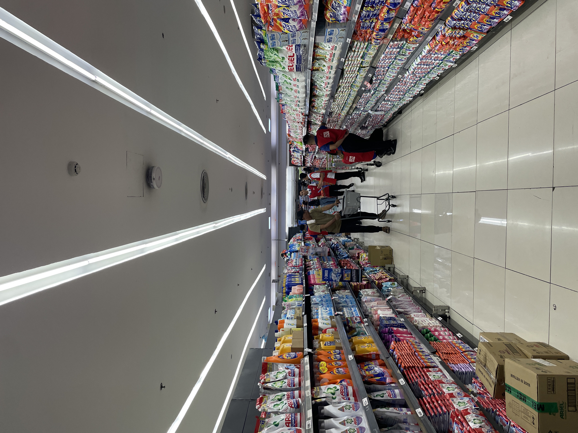The image shows a wide aisle in a supermarket, likely a branch of SM Store or a similar large retail chain, given the prevalence of SM brands and the general store layout. The aisle is flanked by tall shelves stocked with laundry detergents, fabric softeners, and cleaning supplies. Fluorescent lights are mounted in long rows on the ceiling, illuminating the space.
Several people are visible in the aisle. On the left, two shoppers are pushing a shopping cart. Further down the aisle, several store employees are wearing red vests with identification numbers, suggesting they are assisting customers or organizing merchandise. One employee, wearing a red vest labeled "1520," is interacting with the shelves. Another employee in a similar vest is pushing a red trolley. A few other shoppers are seen in the background, also with shopping carts.
The products on the shelves are predominantly in colorful packaging, with brands like Ariel, Tide, Breeze, and Downy clearly visible. The floor is tiled with large, light-colored squares, and there are some cardboard boxes of merchandise stacked on the floor near the entrance of the aisle. The overall impression is of a well-stocked and busy supermarket. The presence of Filipino brands and the general ambiance strongly suggest this location is in Davao City, Philippines.