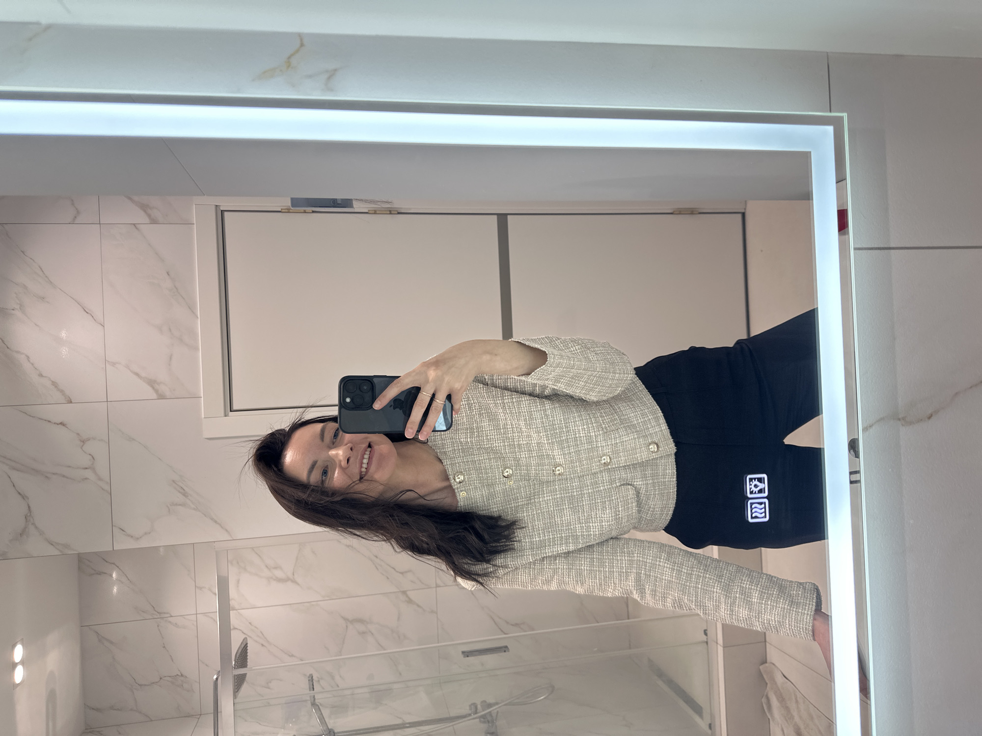 A mirror selfie shot shows a woman standing in a bathroom with marble-like tile walls. She is smiling while holding a smartphone, wearing a light-colored tweed jacket with gold buttons and black pants. The bathroom includes a glass shower door. Location: The city of Прикамский, Russia.