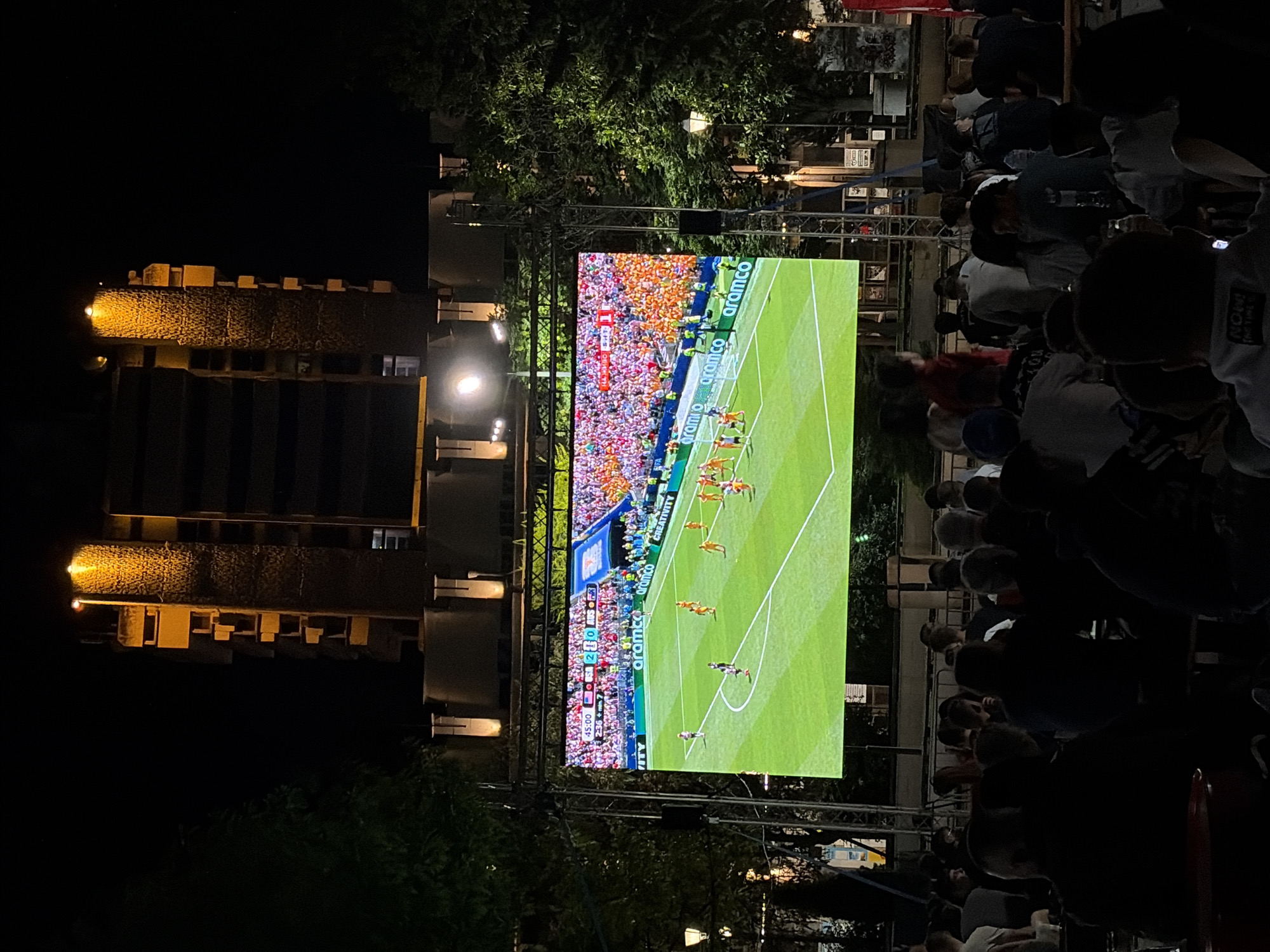 An outdoor night scene shows a large screen displaying a soccer match with a crowd of people watching it. The screen, mounted on a metal scaffolding, shows a soccer field with players and a scoreboard. The scoreboard indicates the score as 2-0. The crowd, silhouetted against the light from the screen and surrounding streetlights, is seated at tables, appearing to be in a relaxed setting like an outdoor bar or fan zone. In the background, a tall, Brutalist-style building with illuminated windows and rough concrete texture looms in the darkness. Trees frame the scene, adding a natural element to the urban environment. The overall atmosphere is that of a public viewing event for a sports game.
