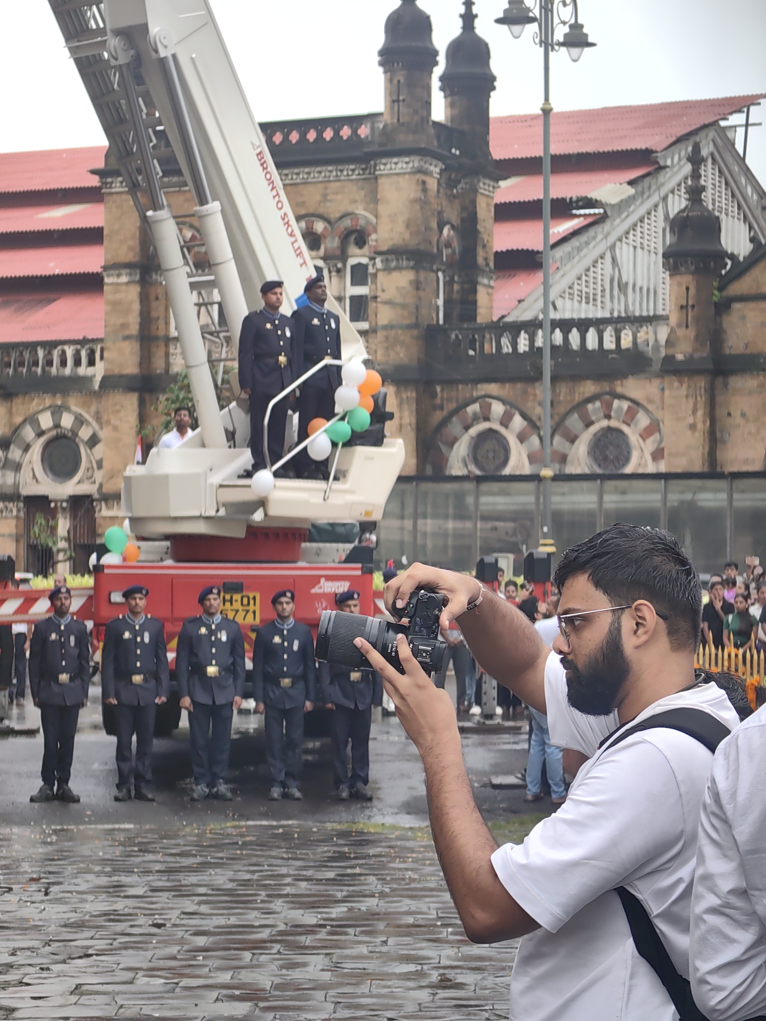 The image captures an event likely occurring at the Chhatrapati Shivaji Maharaj Terminus (CSMT) in Mumbai, India, given the iconic architecture in the background. In the foreground, a man with a beard and glasses is holding a camera and taking a photograph. He is wearing a white t-shirt. Behind him, a group of men in navy blue uniforms with caps stand at attention. These men appear to be part of an official procession or ceremony, possibly firefighters or military personnel, as indicated by their matching attire and organized formation. A large, white aerial ladder platform, identified by the text "BRONTO SKYLIFT," is prominently positioned. Several balloons, colored orange, white, and green, are attached to the platform, suggesting a celebratory or nationalistic event. The ground is wet, indicating recent rain. The background features the distinctive historical architecture of CSMT, characterized by its red-tiled roofs and arched windows.