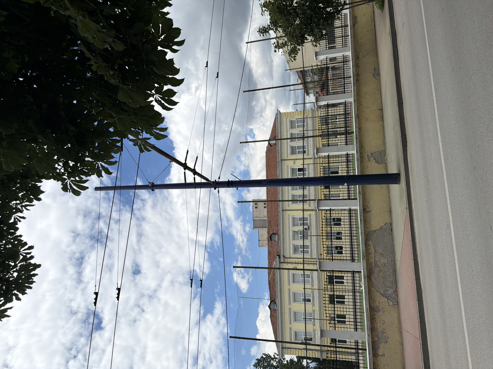 The image shows a street view in Pazardzhik, Bulgaria, facing a yellow building with a red-tiled roof. The building appears to be a school or a public institution, with numerous windows and a fence surrounding the property. Overhead, there are power lines and a tall blue pole, likely a utility pole or part of a public transportation system. The sky is partly cloudy, with a mix of blue sky and white clouds. Trees are visible in the foreground and on the right side of the image, with some leaves obscuring parts of the sky. The street in the foreground is asphalt with white markings. A weathered yellow wall with patches of exposed concrete separates the sidewalk from the building's fence. The overall scene depicts an urban environment on a bright day.
