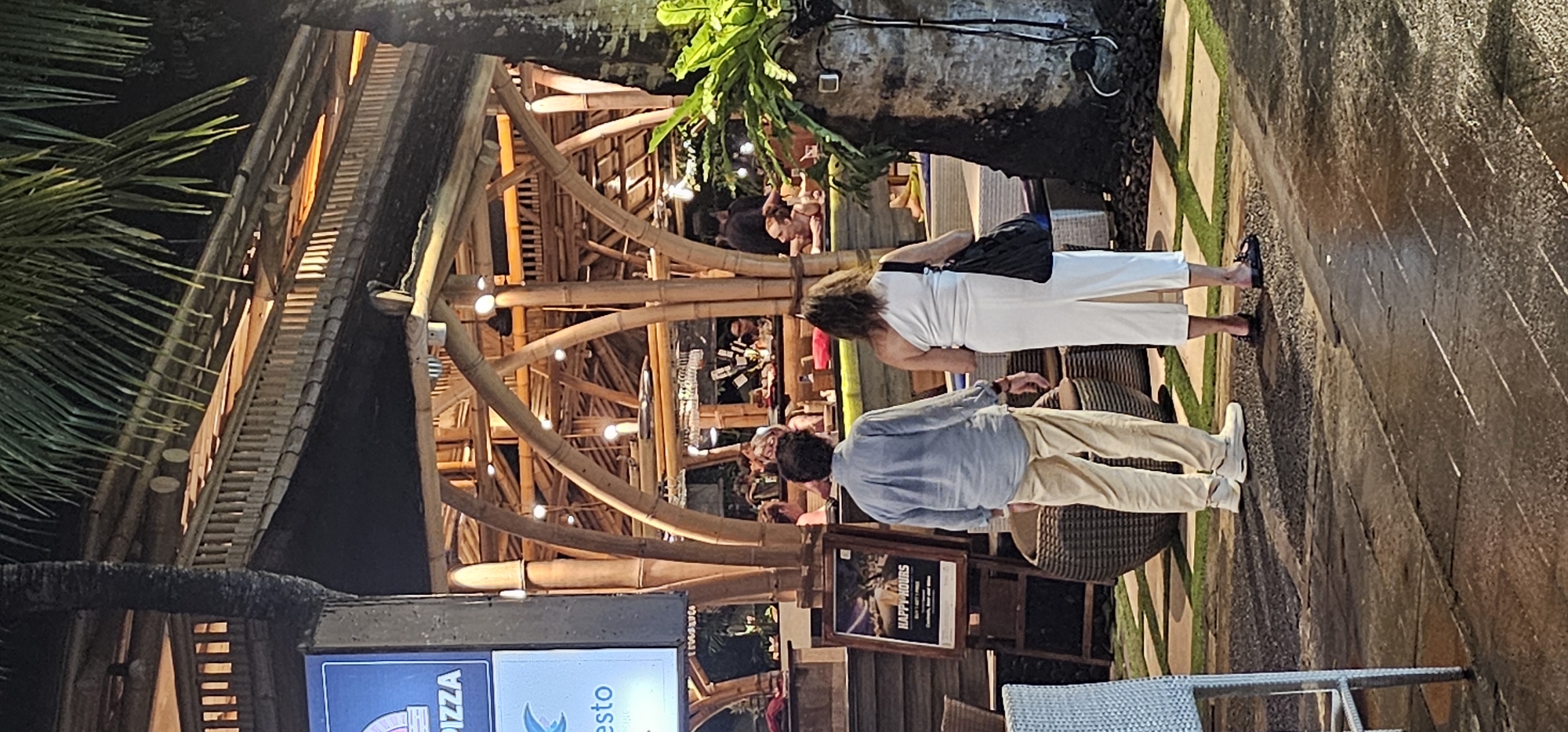 The image captures an evening scene at a tropical-themed outdoor restaurant or bar, consistent with a setting in Denpasar, Indonesia. The main subjects are two people standing in the foreground, with their backs mostly turned to the viewer. On the right, a woman wears a white sleeveless jumpsuit or dress and carries a black shoulder bag. To her left, a man is dressed in a light blue long-sleeved shirt and beige pants, wearing white sneakers. They appear to be patrons either walking through or observing the lively establishment.
The structure of the restaurant is distinctive, built predominantly from numerous bamboo poles and wooden beams, creating an open and airy framework. Small, warm-toned lights are strung along the bamboo, illuminating the space and creating a inviting nighttime ambiance. Lush green palm fronds and other tropical foliage are visible, enhancing the exotic atmosphere.
In the background, various other people are seated at wicker or rattan tables and chairs, dining or socializing. The ground features a pathway of paved stones interspersed with sections of artificial green turf.
Notable details include a small wooden sandwich board sign in the mid-ground displaying "HAPPY HOURS" and "BUY 1 GET 1 FREE". Another partially visible sign on the far left side mentions "PIZZA" and "esto", likely referring to a restaurant or dish. The overall scene suggests a casual and relaxed social gathering during the night, indicative of a resort or tourist-friendly location.