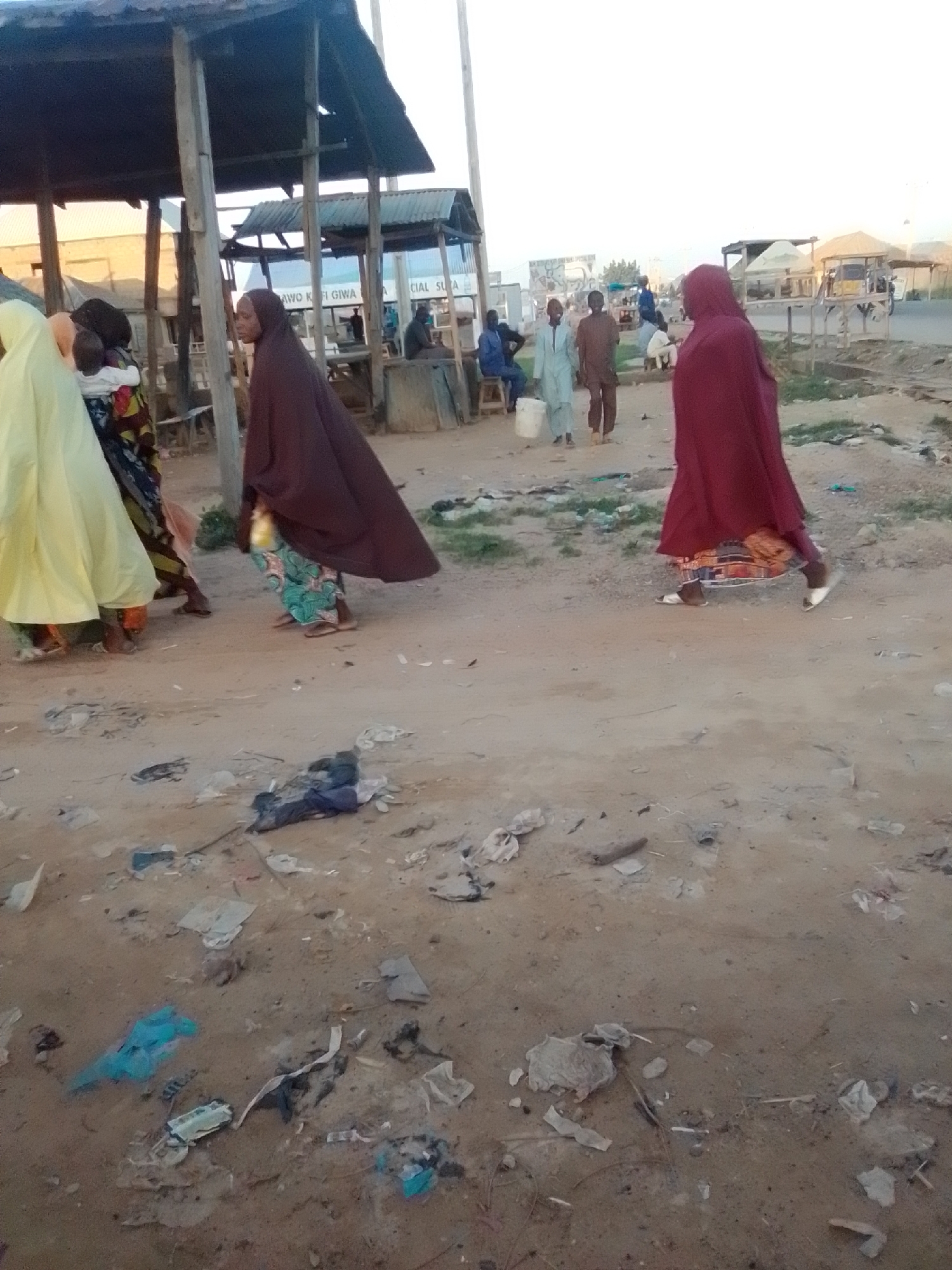 The image depicts a street scene in Gombe, Nigeria, characterized by a dusty, unpaved ground littered with debris, including plastic bags, paper scraps, and discarded items. In the foreground and middle ground, several individuals are present. On the left, three people are partially visible, two of whom are women wearing traditional Islamic attire. One woman is wearing a light yellow hijab and dress, while another is dressed in a dark brown hijab and dress. A third individual, possibly a child, is held by one of the women. In the middle of the scene, a woman in a dark brown hijab and patterned skirt is walking away from the camera. To the right, another woman in a red hijab and dress is also walking away from the camera. In the background, several structures are visible, including market stalls with corrugated metal roofs and wooden supports. Other individuals, presumably men, are seen standing or sitting near the stalls. The overall environment suggests a bustling, informal marketplace or roadside settlement under a bright, possibly overcast sky.