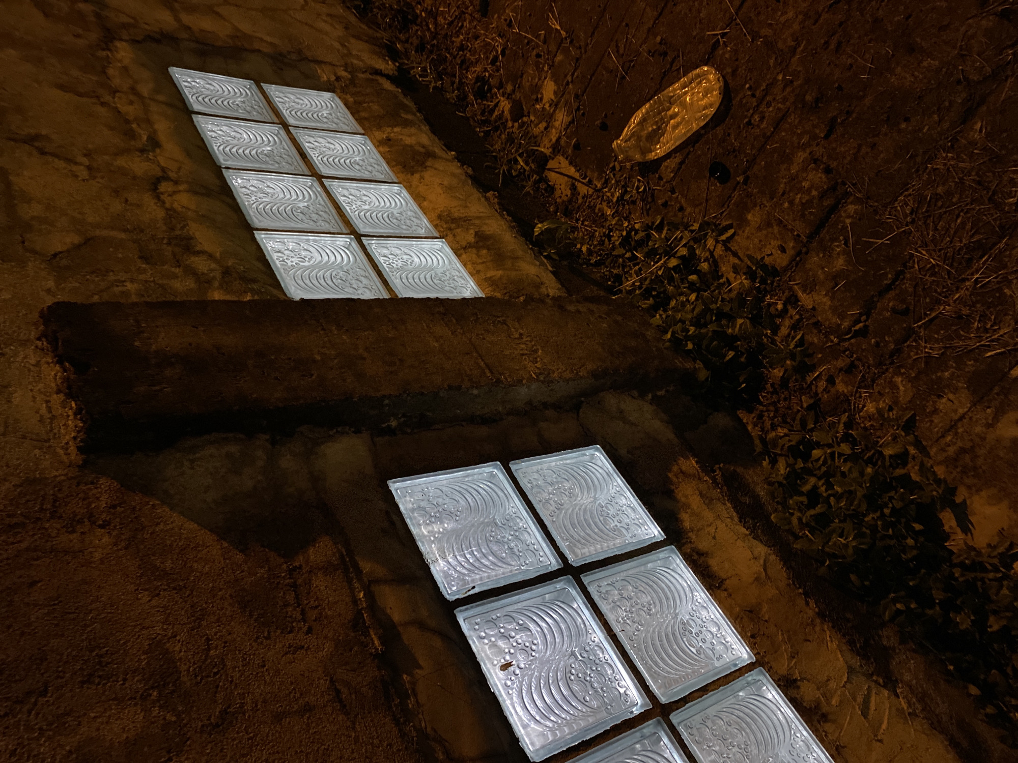 This image depicts an exterior scene at night, likely in an urban or semi-urban environment. The foreground shows a wooden or concrete surface covered with dry leaves, twigs, and some sparse vegetation. A discarded clear plastic bottle with a yellow-orange tint lies on this surface.
Dominating the scene are two sets of glass block windows set into a rough, textured concrete wall. The window on the left consists of four glass blocks arranged in a 2x2 grid. The window on the right is larger, featuring eight glass blocks in a 2x4 grid. The glass blocks have a textured, wave-like pattern and are illuminated from within, casting a soft, white light.
A vertical concrete pillar separates the two window sections. The overall lighting suggests artificial illumination from the windows and possibly ambient streetlights, creating strong shadows. There are no people or animals visible in the image. The scene appears to be a back alley or a service area of a building. The location is specified as Davao City, Philippines.