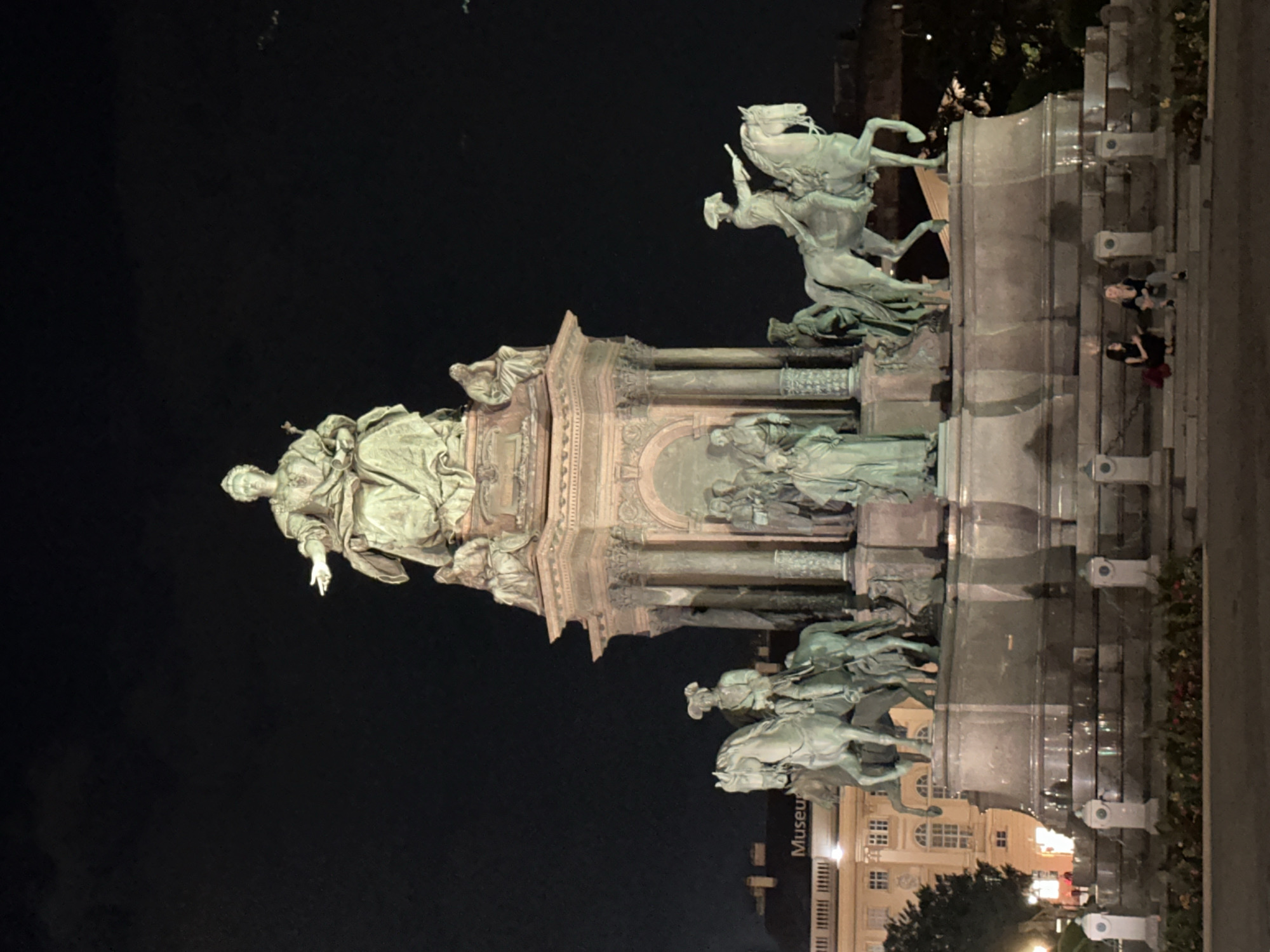 The image shows the Maria-Theresien-Denkmal in Vienna, Austria, at night. The monument features a large statue of Empress Maria Theresa seated on a throne, surrounded by statues of other figures and equestrian figures. Two women are seated on the steps of the monument in the foreground, with one woman holding a bag. The monument is illuminated by spotlights, casting a dramatic glow on the bronze sculptures. In the background, buildings and trees are visible, with the dark night sky above. The scene appears calm and quiet, with the monument serving as a majestic centerpiece in the dimly lit square.