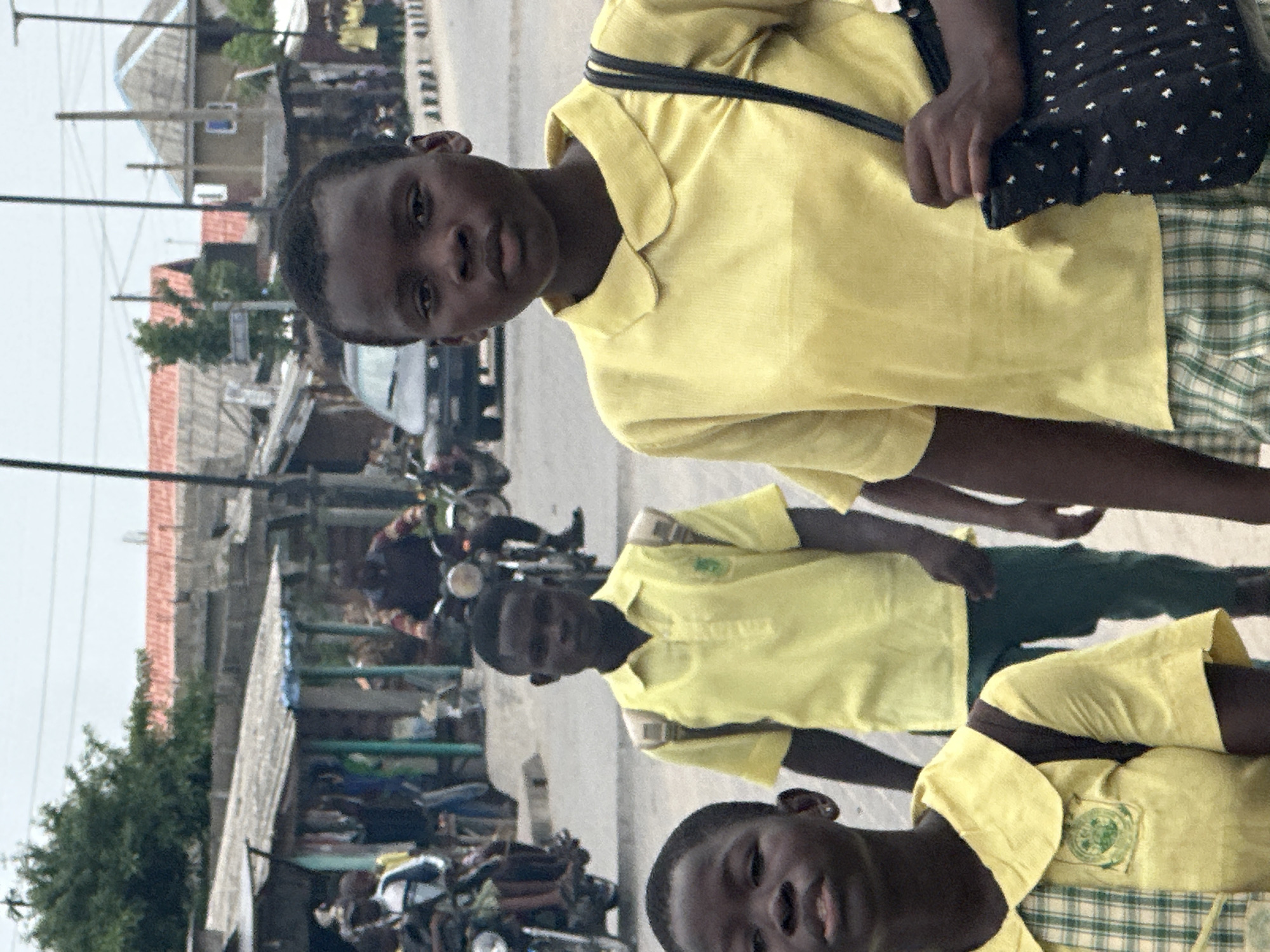 The image captures three young individuals, likely students, standing on a street in Badagry, Nigeria, during daytime. The photograph is oriented vertically, showing the subjects diagonally from the top-left to the bottom-right.
The person closest to the camera is on the left, looking directly at the viewer with a neutral expression. They are wearing a yellow polo shirt, a green and white plaid skirt or lower garment, and carry a dark shoulder bag with white polka dots. Behind them, a second person, also in a yellow polo shirt and green lower garment, looks towards the camera. A circular green school emblem is visible on their left chest. Further back and to the right, the third individual, partially visible, also wears a yellow polo shirt with a similar plaid skirt and school emblem, and looks towards the camera.
The setting appears to be a bustling street or thoroughfare. In the background, a row of buildings with corrugated metal roofs and unpainted walls line the road. Several motorcycles are visible, some parked, others possibly in motion, along with a partially obscured car. Utility poles and overhead wires crisscross the frame. The lighting is bright and even, indicating daylight, possibly under an overcast sky, as there are no harsh shadows. The overall scene reflects a common street environment in a West African town. No specific text is clearly legible on signs or buildings, but the school emblems on the uniforms suggest an educational context.
