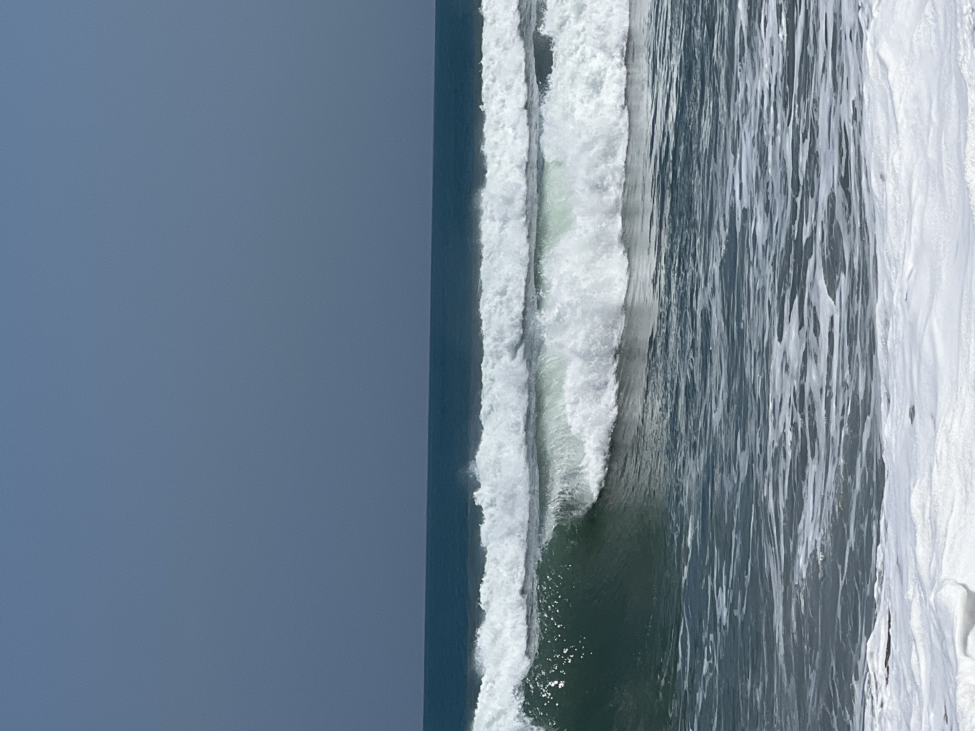 The image depicts a serene seascape with two distinct waves cresting in the foreground. The ocean is a deep teal, transitioning to a lighter, foamy white where the waves break. The sky above is a muted, cloudless blue, creating a stark contrast with the turbulent water. This scene is likely located on the coast of Kuta, Indonesia, a popular surfing destination known for its dynamic waves and expansive beaches. There are no people or man-made objects visible in the frame, emphasizing the natural beauty of the ocean and sky.