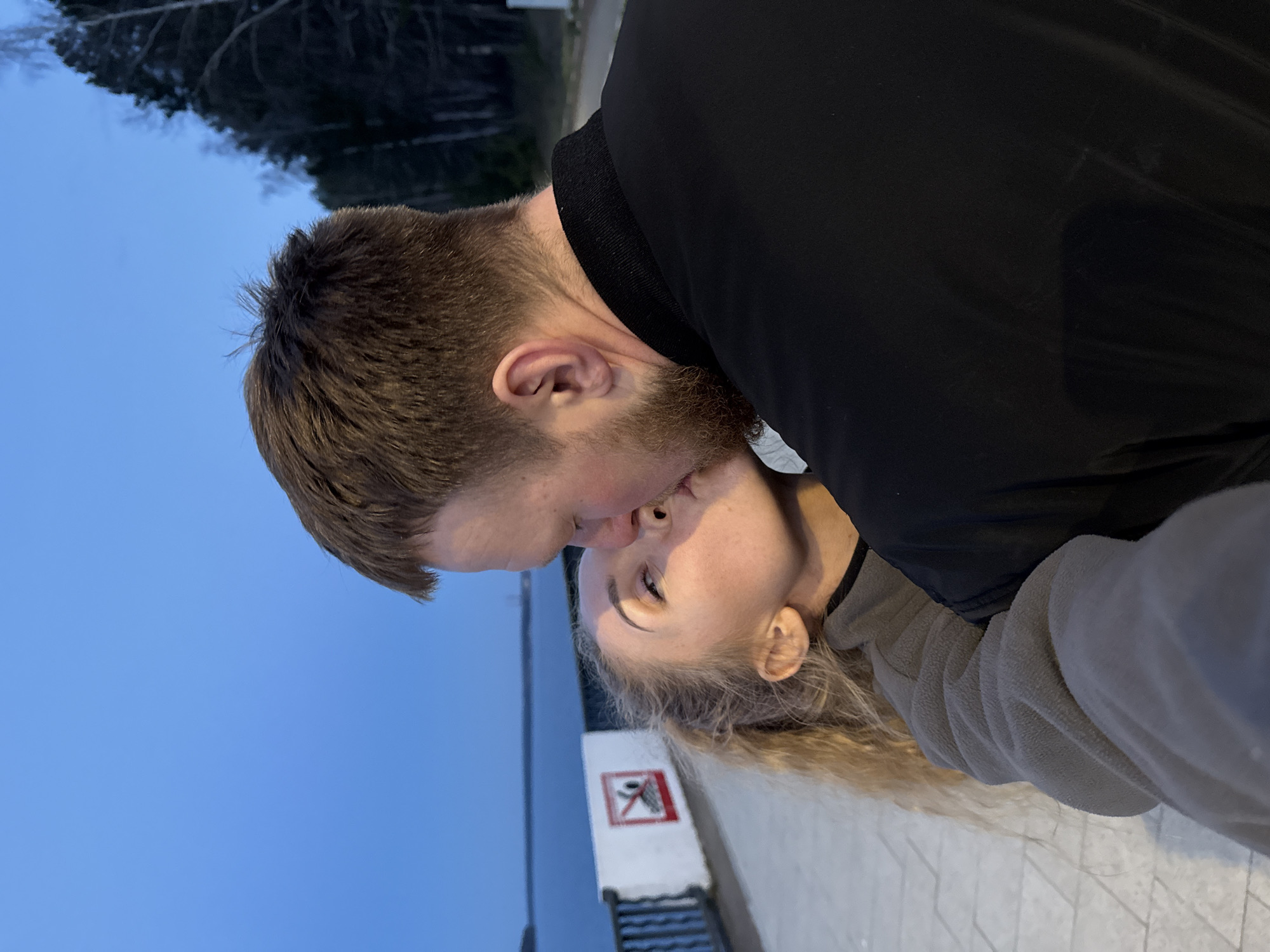 Two people are kissing. The person on the left has short brown hair and a beard and is wearing a black jacket. The person on the right has long blond hair and is wearing a gray fleece jacket. They are outside, with a body of water in the background. A sign is visible in the background. The location is likely in the city of Chaikovsky, Russia.