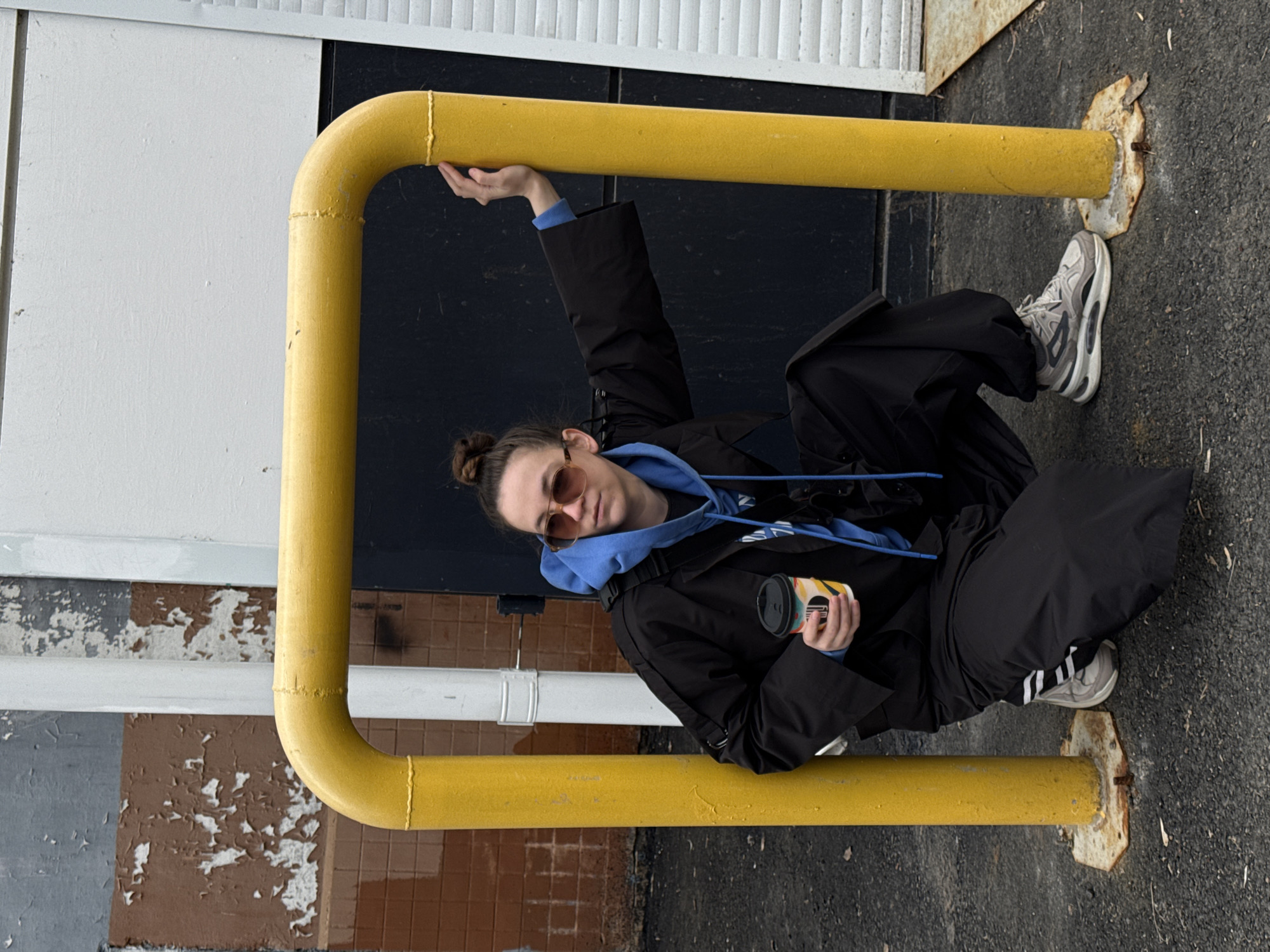 A woman is posed in an urban setting. She is leaning on a yellow, rectangular, metal barrier that is secured to the ground. The barrier is positioned in front of a building facade with sections of white, gray, black, and brown panels with peeling paint. The surface beneath the barrier is dark gray asphalt. The woman has a bun hairstyle and is wearing sunglasses, a light blue hooded sweatshirt, and a black jacket. She is holding a disposable coffee cup in her right hand and has one arm raised, holding onto the yellow barrier. Her legs are bent and she is wearing oversized black pants and white sneakers with black stripes. The location is Chaikovsky, Russia.