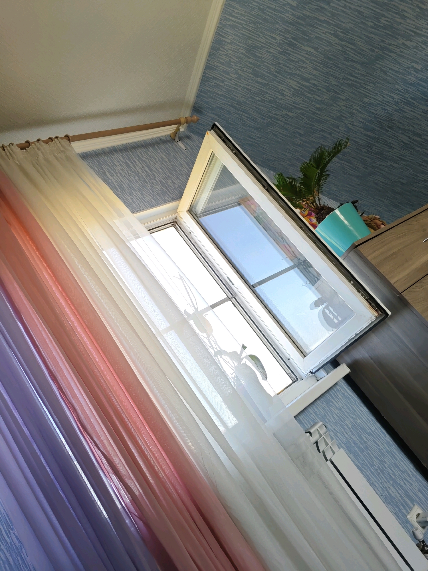 The image depicts an interior scene, likely a room, with a window partially open. A sheer curtain, featuring a gradient of colors from purple to pink to white, hangs to the left of the window. The window itself is white-framed and reflects the blue sky. Outside the window, a bright sky is visible, with hints of green foliage. On a shelf to the right of the window, a potted plant with green leaves sits next to a turquoise pot. A wooden cabinet or dresser is partially visible on the far right. The walls are covered with blue, textured wallpaper. A white radiator is positioned beneath the window. The perspective is from a low angle, looking upwards towards the window and ceiling. There are no people or animals present in the image, nor any indication of the specific location within Rostov-on-Don, Russia.