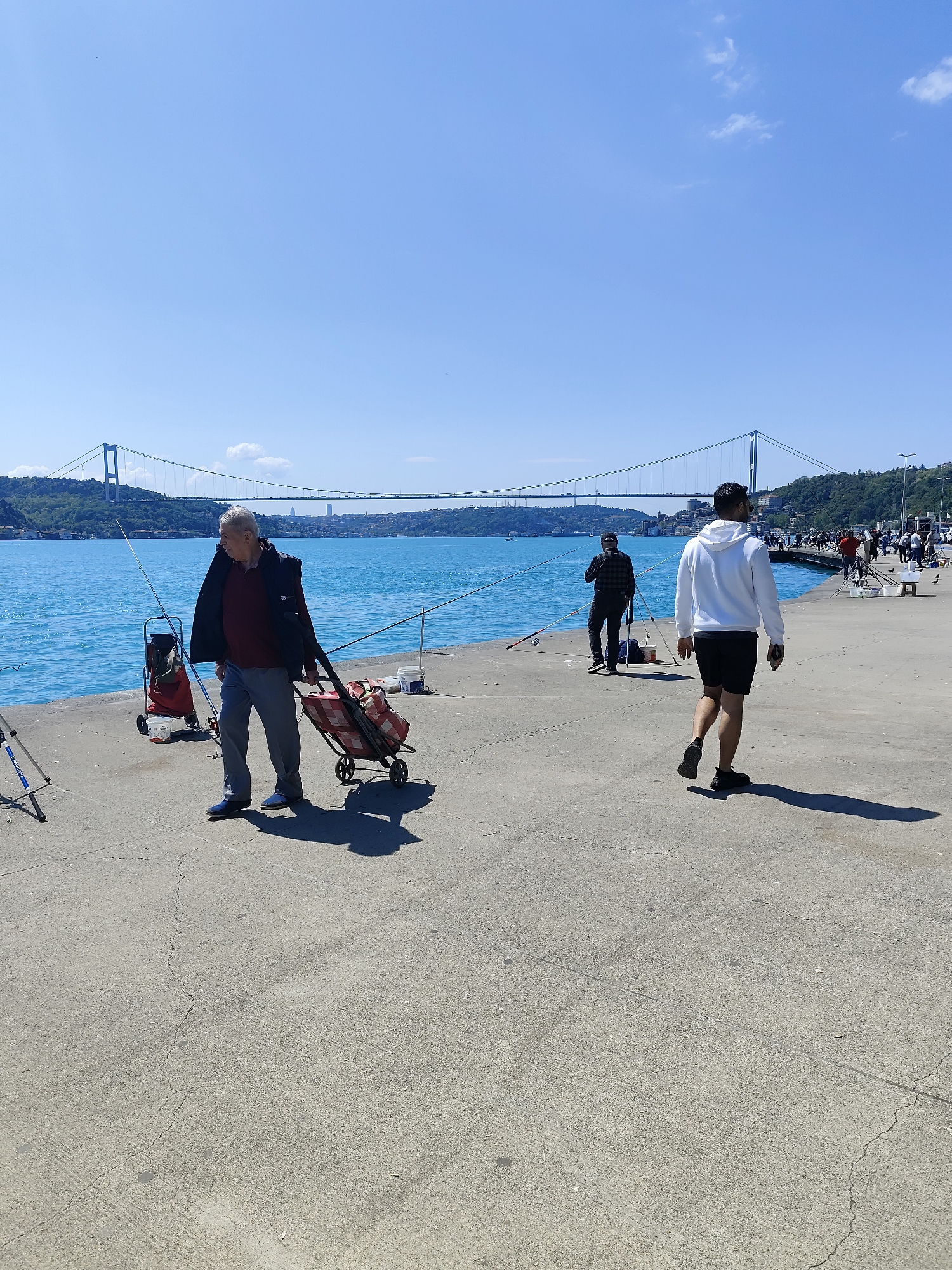 The image depicts a daytime scene in Sarıyer, Turkey, with the Bosphorus Bridge in the background. The foreground features a concrete pier where several people are fishing. An elderly man wearing grey trousers, a maroon shirt, and a black jacket is hauling a red shopping cart. Fishing rods are set up along the pier's edge. A man wearing a white hoodie and black shorts is walking away from the camera. Other people are fishing further down the pier, near a crowd, with various fishing equipment and supplies nearby. The water is blue, and the sky is clear with a few clouds. Shadows from the people and objects are visible on the pier.