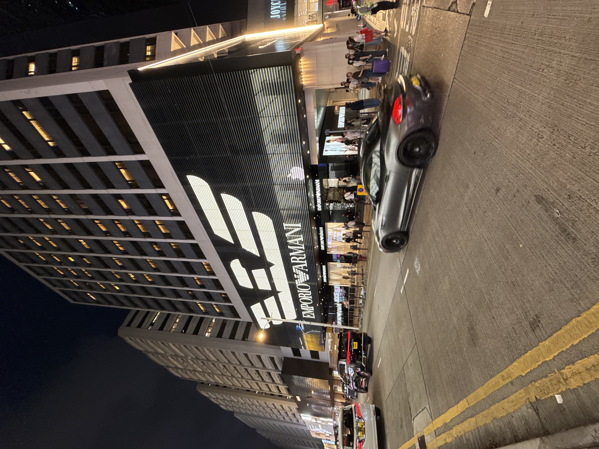 Captured at night, the image presents a tilted view of a city street featuring an Emporio Armani store. The store's prominent signage, adorned with the brand's logo and name in large, illuminated letters, is a focal point. People are visible in front of the store and on the sidewalk, suggesting pedestrian activity. Several vehicles, including cars and vans, line the street, indicating urban traffic. Tall buildings, with illuminated windows, frame the scene, characteristic of a dense urban environment. The street is marked with yellow lines, typical of road markings. Given the presence of the Emporio Armani store and the overall urban landscape, the location is likely in Hong Kong, China.