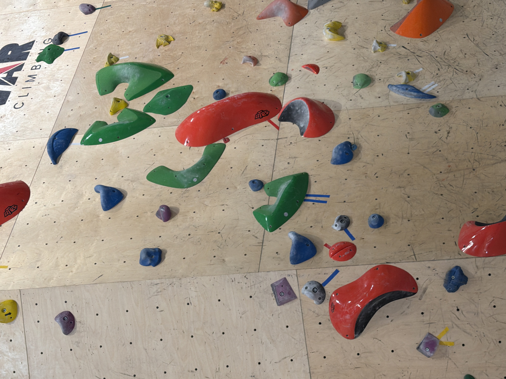 The image shows an indoor climbing wall. The wall is constructed from light-colored plywood and features numerous climbing holds of various shapes, sizes, and colors, including red, green, orange, blue, purple, and yellow. Some holds are marked with tape. The climbing wall has "AR Climbing" written at the top left. The specific location in Yerevan, Armenia is unknown.