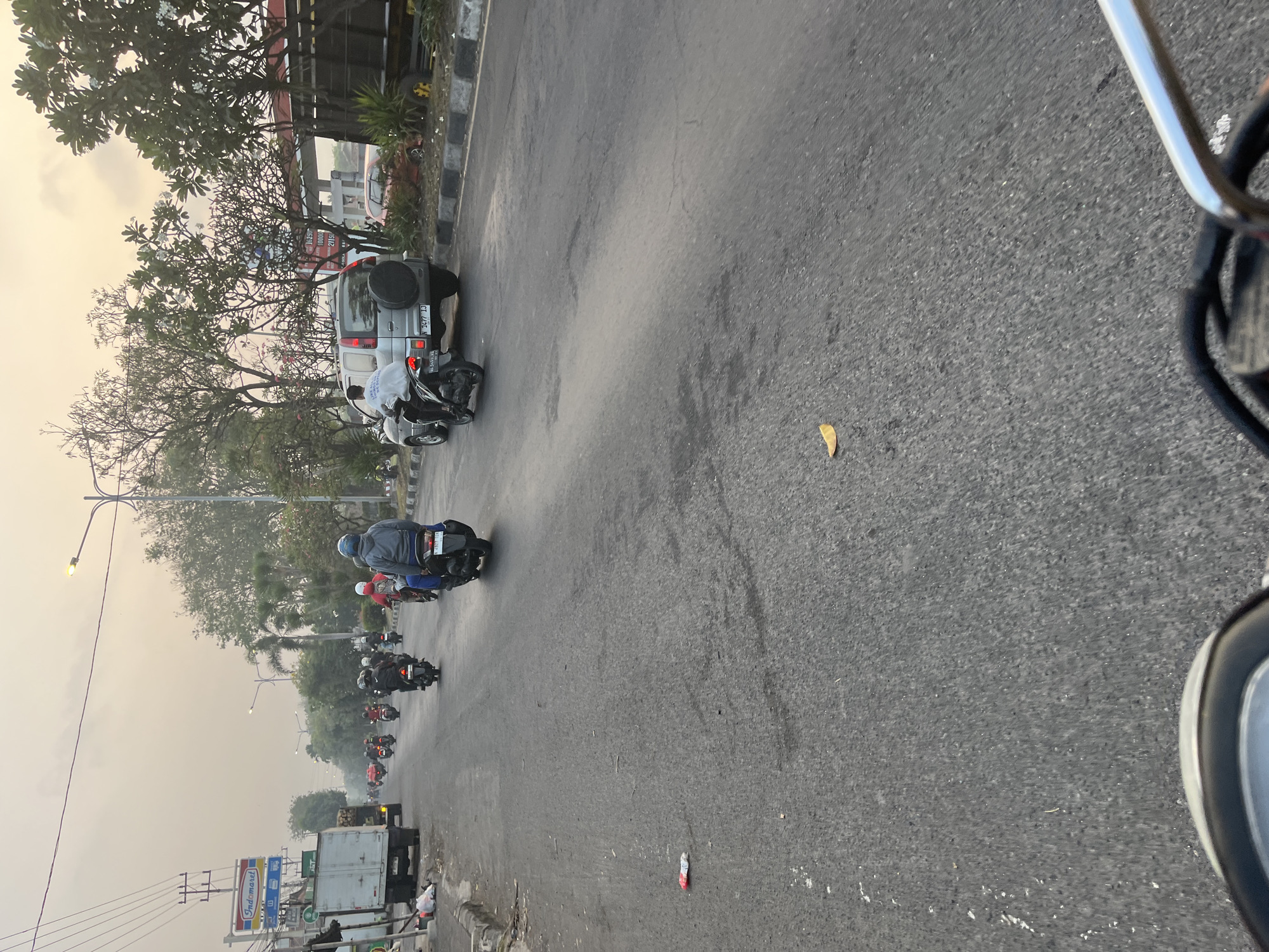The image depicts a street scene in Kludan, Indonesia, likely during the early morning or late afternoon due to the soft, hazy lighting. The road is paved and appears well-trafficked by motorcycles. In the foreground, the handlebars and part of the dashboard of a motorcycle are visible, suggesting the photo was taken from the perspective of a rider.
Several motorcycles are in motion, with riders wearing helmets and some with backpacks. One rider in the middle distance is wearing a blue helmet and a gray jacket. To the right of the road, a silver SUV with a spare tire mounted on the back is parked. Further down the road, a white truck is also visible. The left side of the road features sidewalks, power lines, and a sign for "Indomaret," a convenience store chain common in Indonesia. Trees line the right side of the road, and a gas station canopy is partially visible in the background. The overall impression is of a typical urban or suburban street scene in Indonesia, with everyday traffic and commerce.