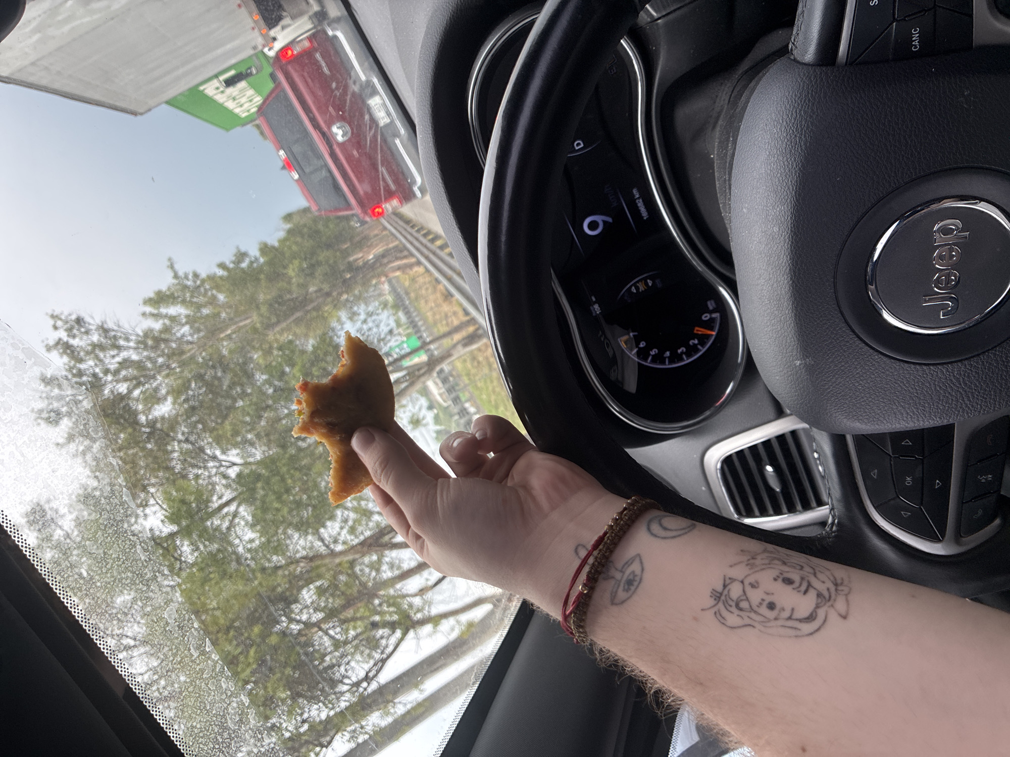 The image, taken from inside a Jeep, shows a person holding a partially eaten pastry in their hand. The steering wheel with the Jeep logo is visible. The dashboard displays a speed of 6 km/h and a mileage of 160,682 km. Outside the vehicle, the environment suggests a highway setting, with a truck visible in the rearview mirror and trees lining the road. The dashboard includes controls for audio, telephone, and vehicle functions. The person's arm is visible, adorned with a beaded bracelet and tattoos. Based on the vegetation and the fact the dashboard is in Spanish, the image could be somewhere in Mexico. It is unlikely to be Tepotzotlán unless there is a highway going through that location.
