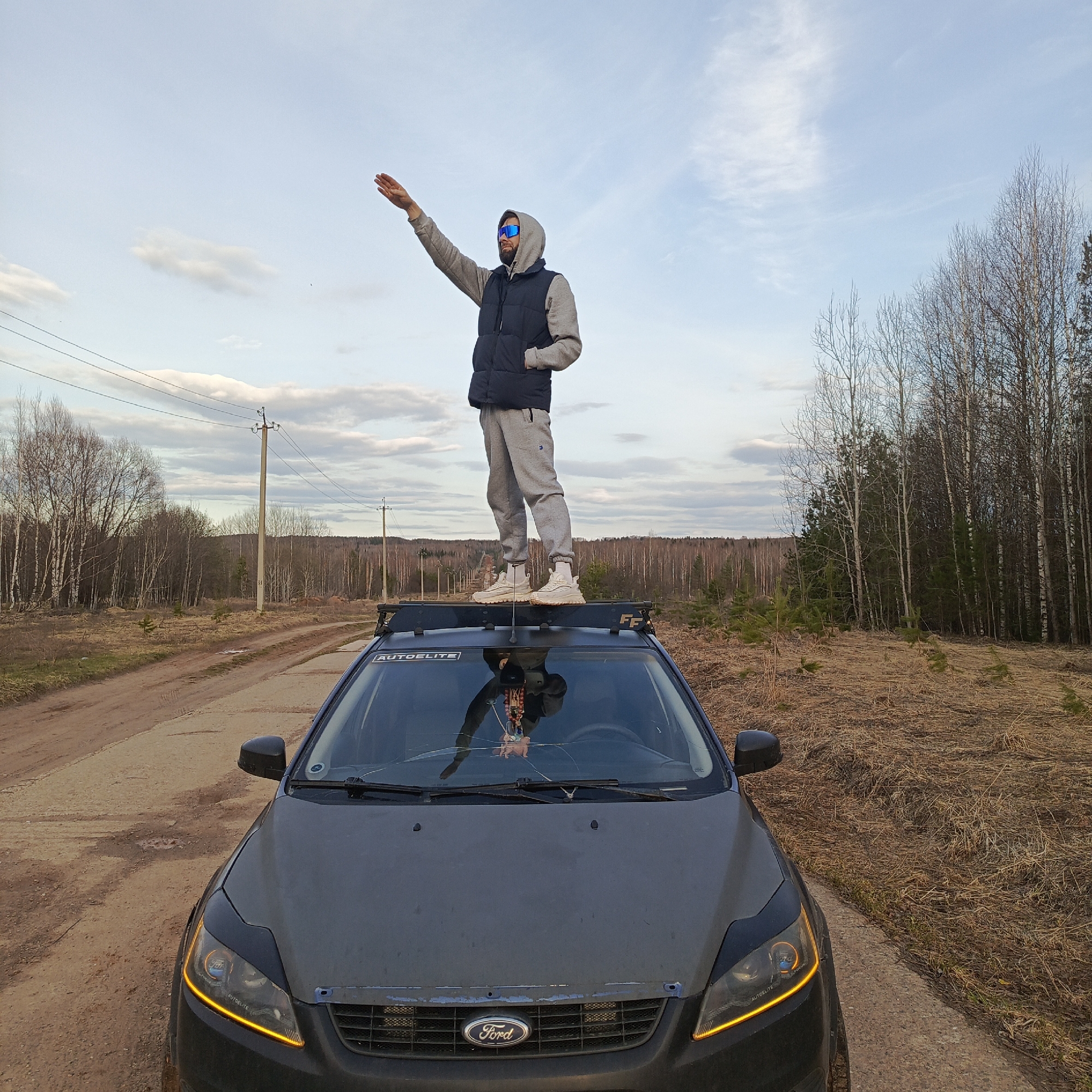 A man stands on the roof rack of a grey Ford Focus car on a dirt road in Марковский, Russia. He is wearing a grey hooded sweatshirt, a black vest, grey sweatpants, and tan sneakers, with blue sunglasses. He is raising his right arm forward in a straight line, palm down. The car has a cracked windshield and a roof rack with the word "AUTOELITE" on it. The background consists of a dirt road, bare trees, power lines, and a cloudy sky. The ground is covered in dry grass.