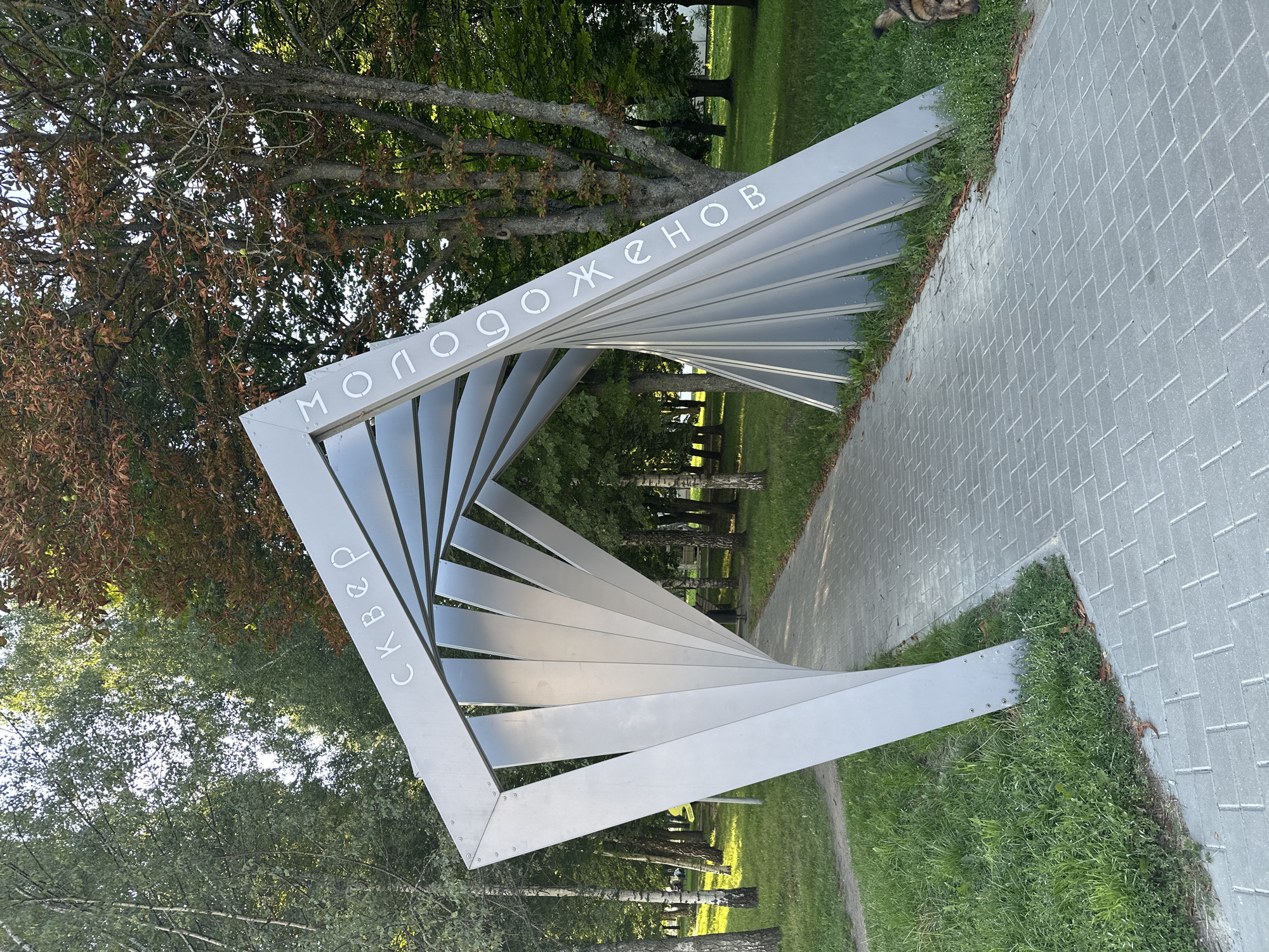 The image shows a modern sculpture entrance in a park with a paved walkway. The sculpture is made of metal and has a twisting, abstract design. Russian Cyrillic text is inscribed on the sculpture, reading "СКВЕР МОЛОДОЖЕНОВ" (SKVER MOLODOZHENOV), which translates to "Youth Park". The park is filled with trees, and the ground is covered with grass and fallen leaves. The sky is visible through the trees and the sculpture. In the lower right corner of the image, a dog is partially visible. The overall impression is of a public park area in an urban setting, likely in a location where Russian or Belarusian is spoken, given the Cyrillic text.