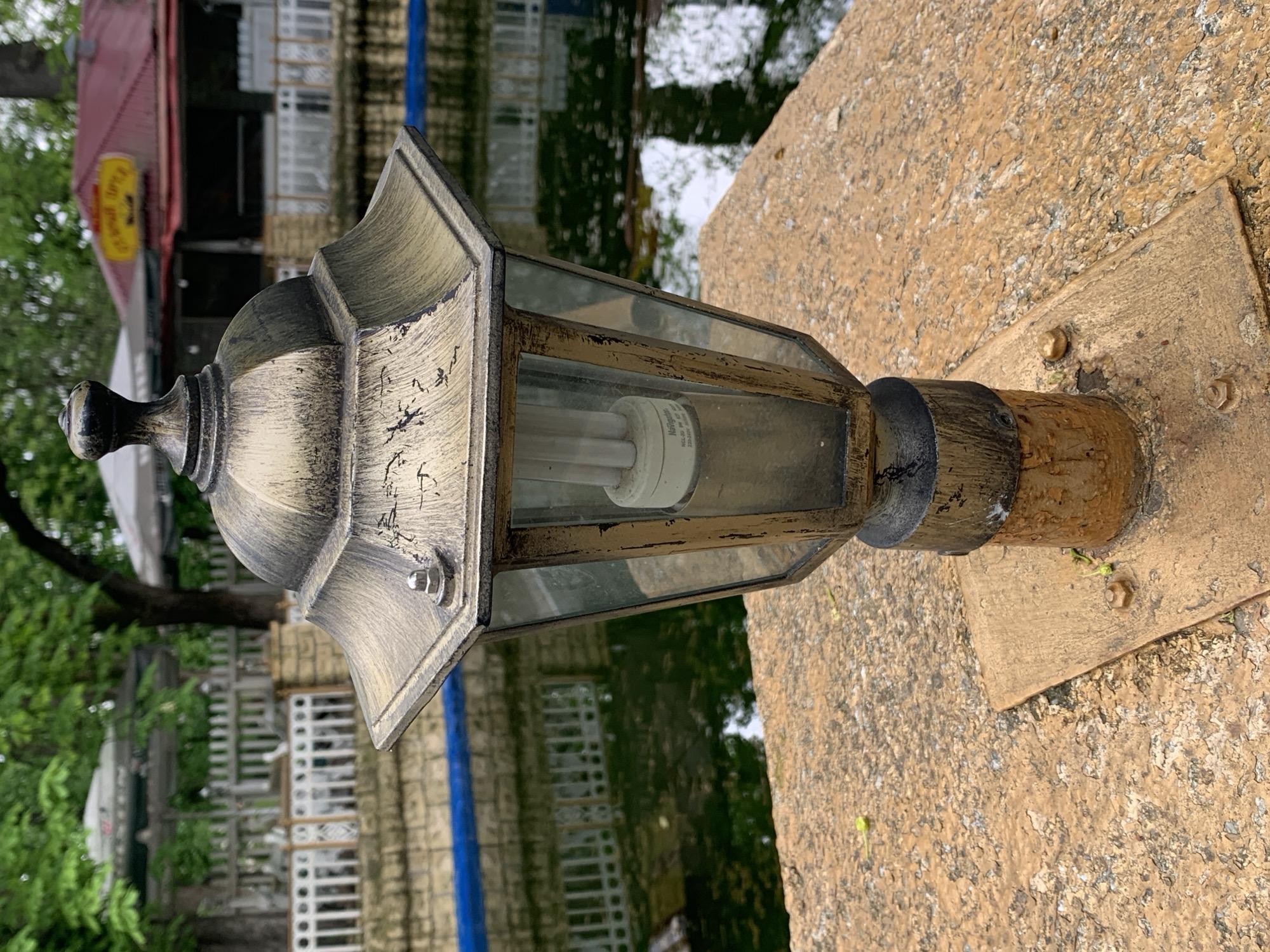 A close-up shot, taken outdoors in daylight, focuses on an ornate, antique-style light fixture. The light is secured to a cracked, weathered concrete surface with a square metal base, fastened with visible bolts. The fixture has a gray and gold color scheme with a clear glass enclosure around a compact fluorescent light bulb. In the background, slightly out of focus, there is a body of water reflecting buildings and greenery. A red roof is visible beyond the water. The scene is located in Stavropol, Russia.