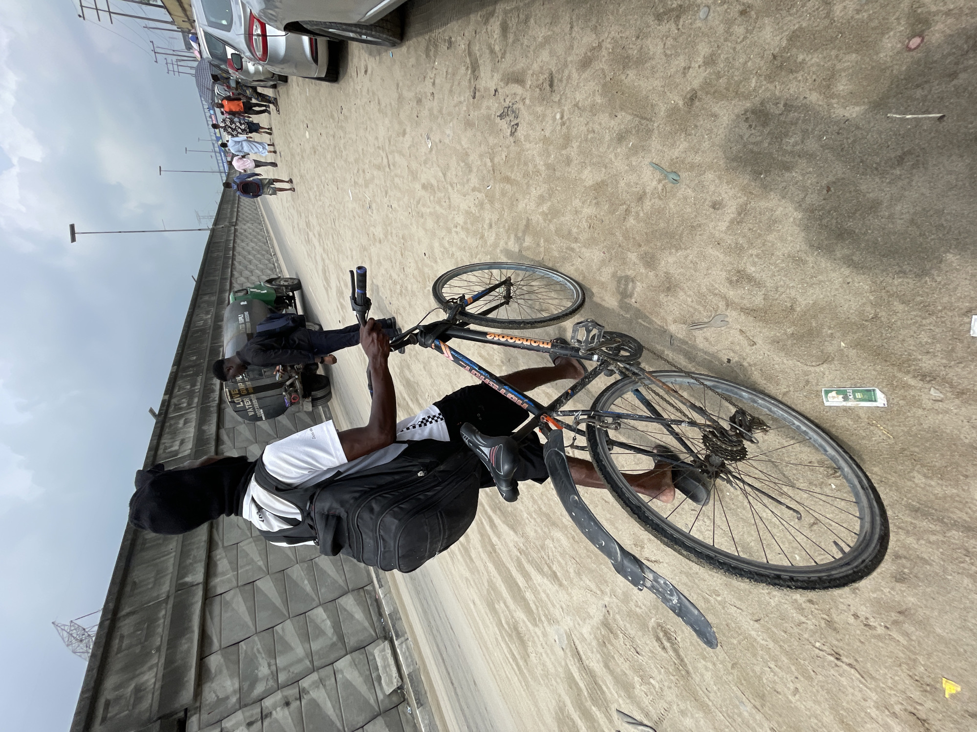 A young man wearing a black head covering, a white t-shirt, and black shorts rides a bicycle on a dirt road in Lekki, Nigeria. He has a black backpack on. The bicycle is black and has orange lettering. In the background, a gray concrete wall runs along the left side of the road. A green water tanker truck is parked behind the man. Several people walk along the road in the distance. Cars are parked on the right side of the road. The sky is cloudy.