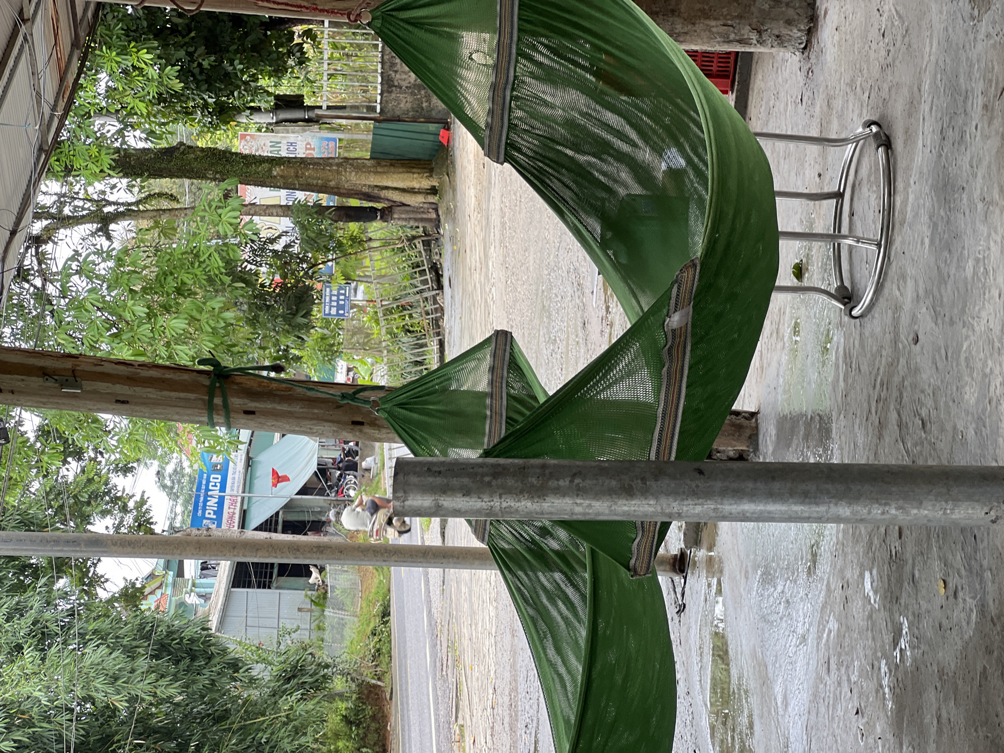 In Thái Sơn Commune, Vietnam, two green mesh hammocks hang suspended from wooden poles and metal frames in an outdoor covered area. The hammocks are empty, with one showing a glimpse of a person's legs within. The ground is a wet, concrete surface, reflecting the overcast sky and surrounding environment. In the background, a road is visible with a person riding a scooter. Beyond the road, buildings with corrugated metal roofs and blue signage are present, surrounded by lush green trees. The scene appears to be a roadside rest area or a small business establishment.